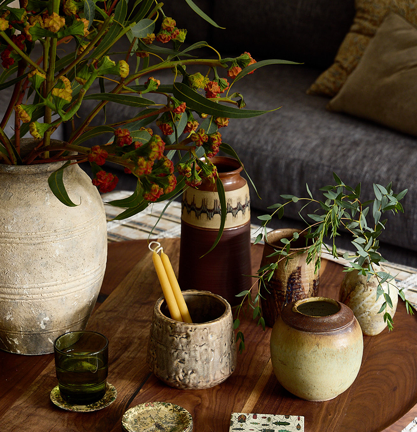 A wooden table with various pottery vases and containers holding plants and candles. A large beige vase contains a leafy green plant with red and yellow flowers. Other smaller vases hold green leafy branches. There is a glass with a dark beverage and a small decorative plate on the table. In the background, part of a sofa with beige and gray cushions is visible.
