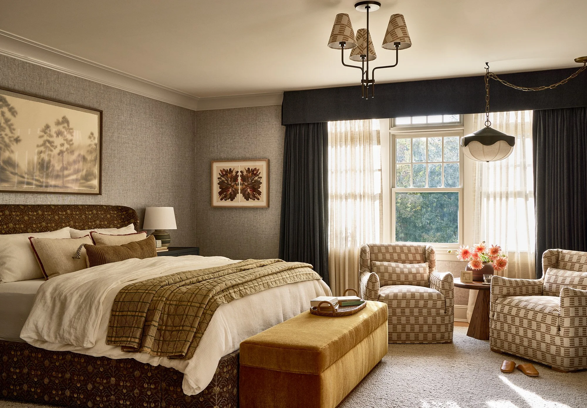 A well-lit hotel room with a large bed, patterned headboard, white linens, and multiple pillows. To the left of the bed is a nightstand with a lamp. There are framed pictures on the textured wallpaper wall. Near the window are two armchairs with chec