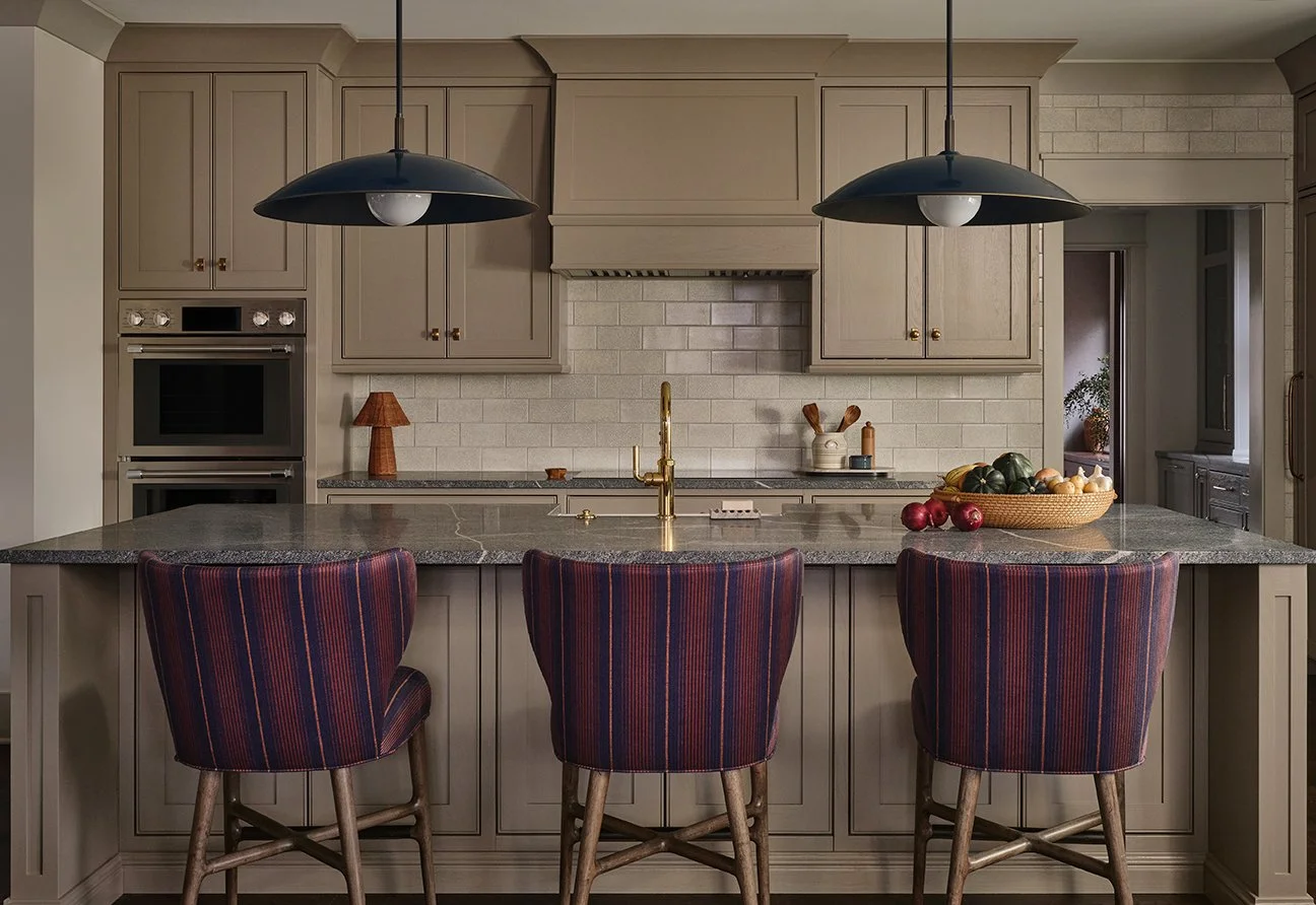 A modern kitchen with beige cabinets, a gray granite island, and two black pendant lights. There are three striped chairs at the island, a bowl of vegetables, and a beige tiled backsplash.