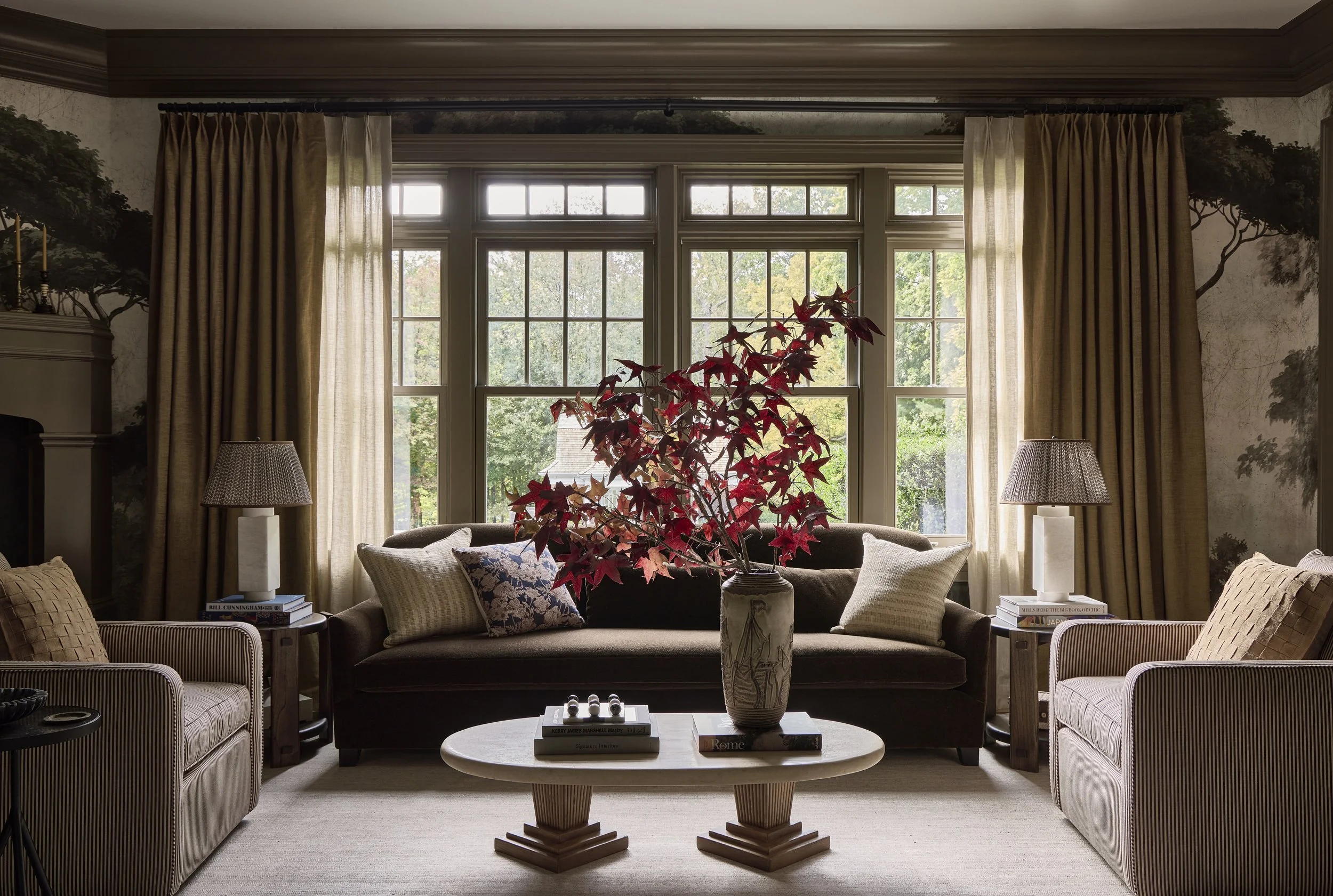 Living room with large window, beige curtains, central vase with red leaves on coffee table, brown sofas, armchairs, side tables, and table lamps.