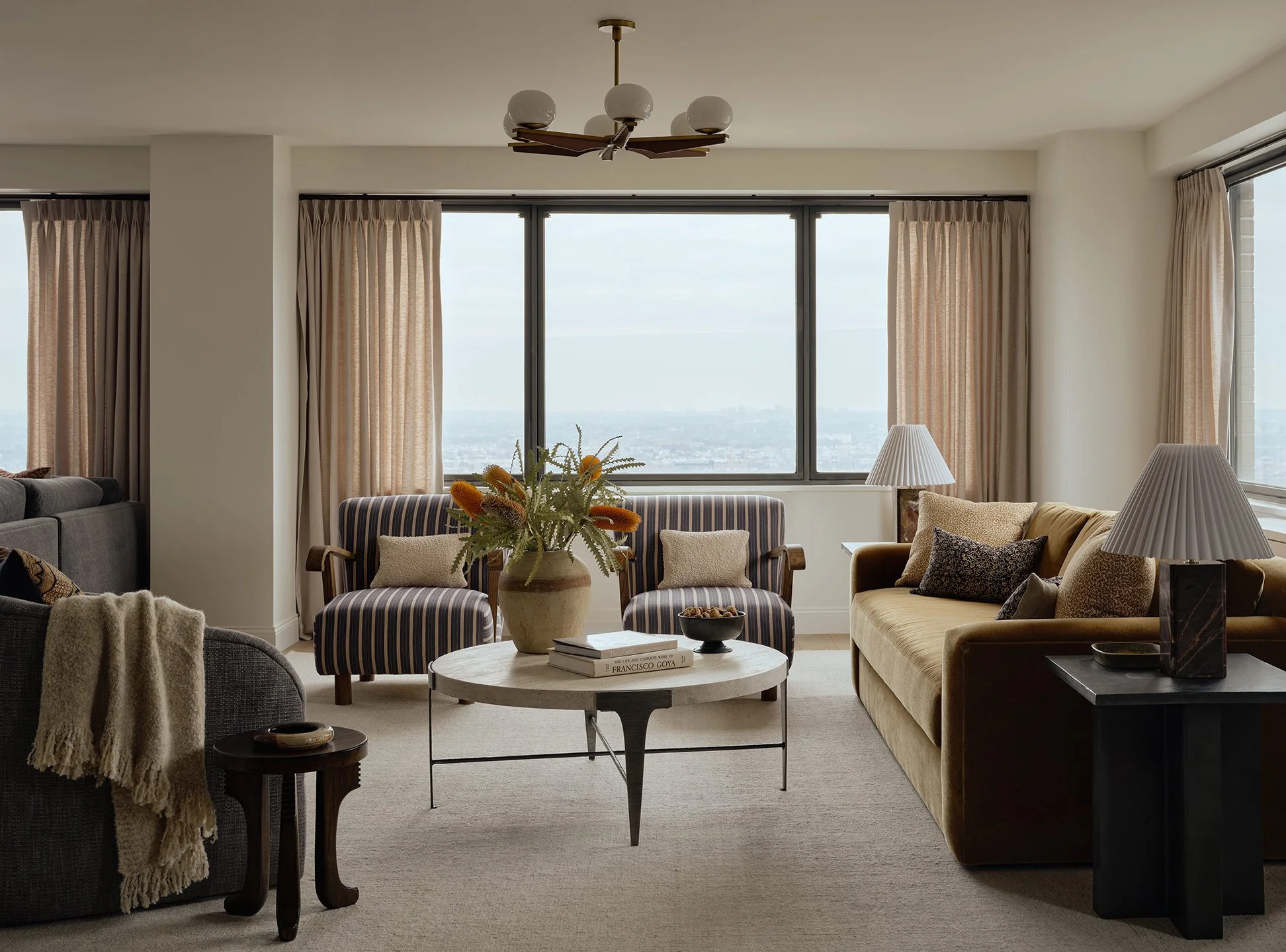 Living room with large windows, beige curtains, a striped armchair, yellow sofa with pillows, a coffee table with a potted plant, lamps, and a view of the city skyline.