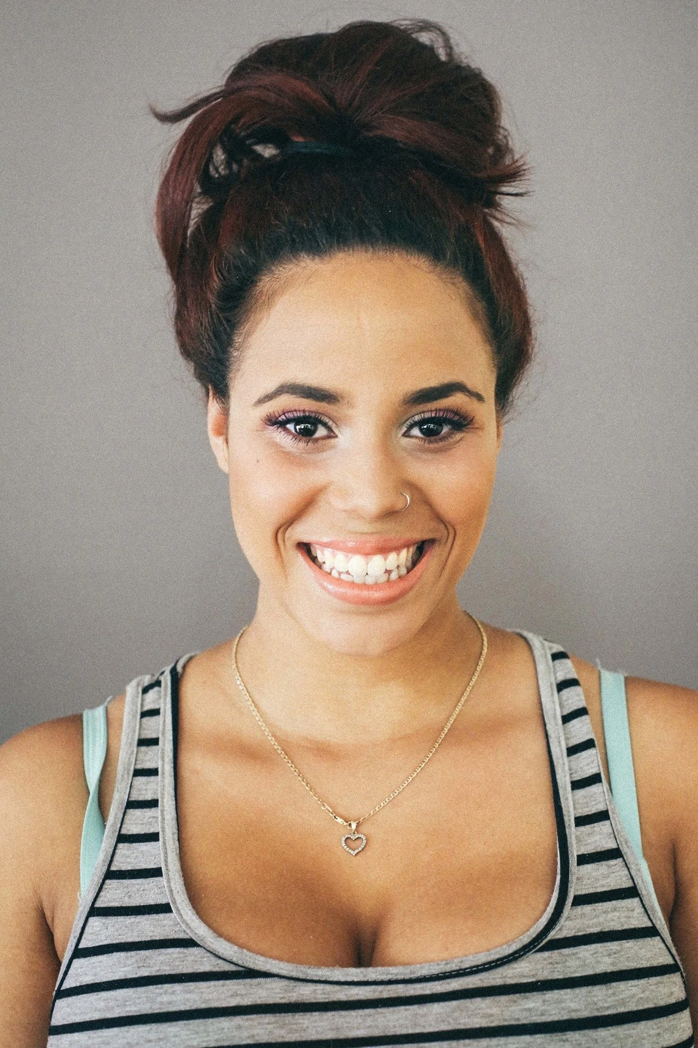A young woman smiling at the camera, wearing a striped tank top, with an updo hairstyle, and a heart-shaped necklace.