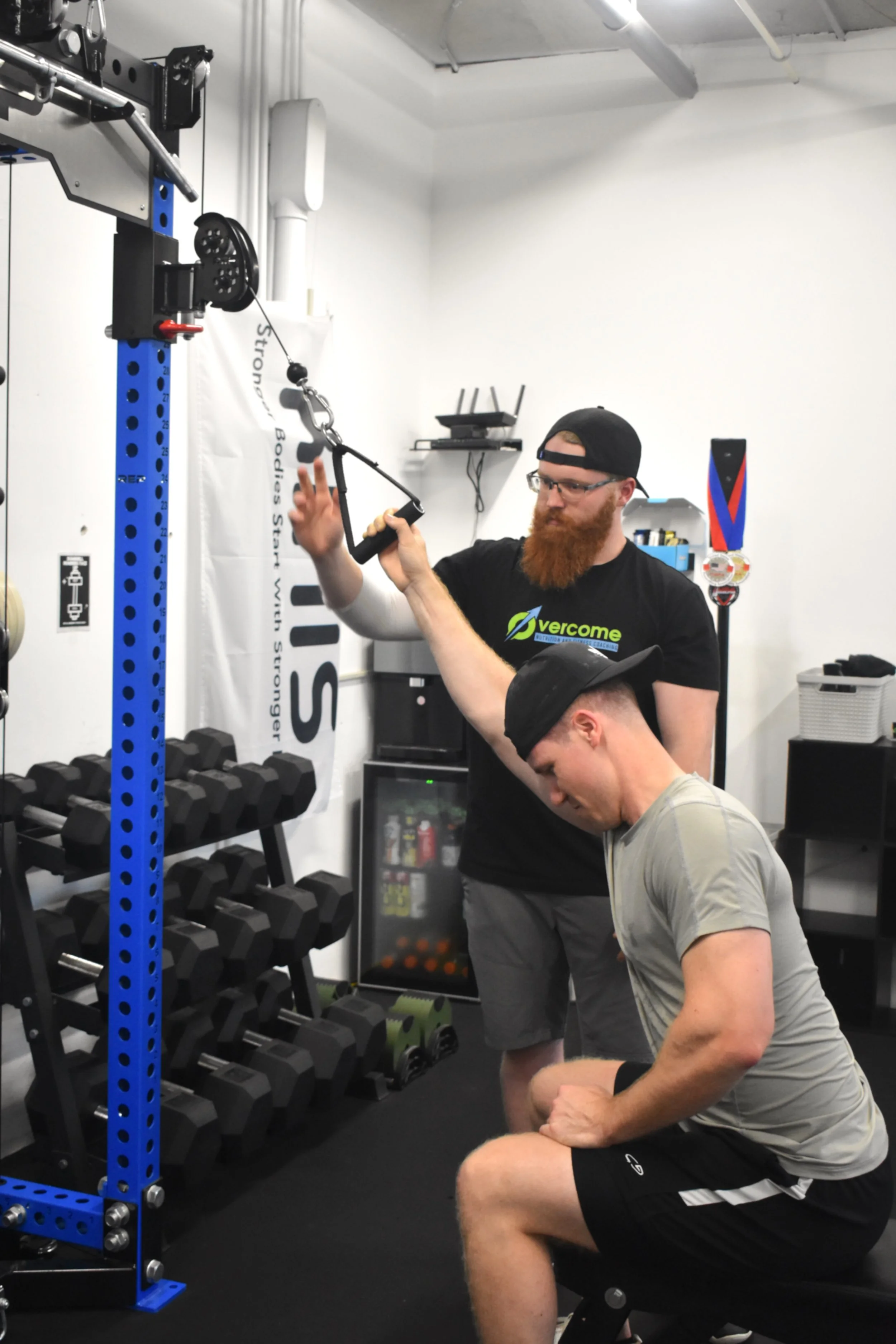A personal trainer assisting a man during a tricep cable pushdown exercise in a gym. The trainer is standing, guiding the exercise, while the man is kneeling, gripping the cable handle, wearing a cap, gray t-shirt, and black shorts.
