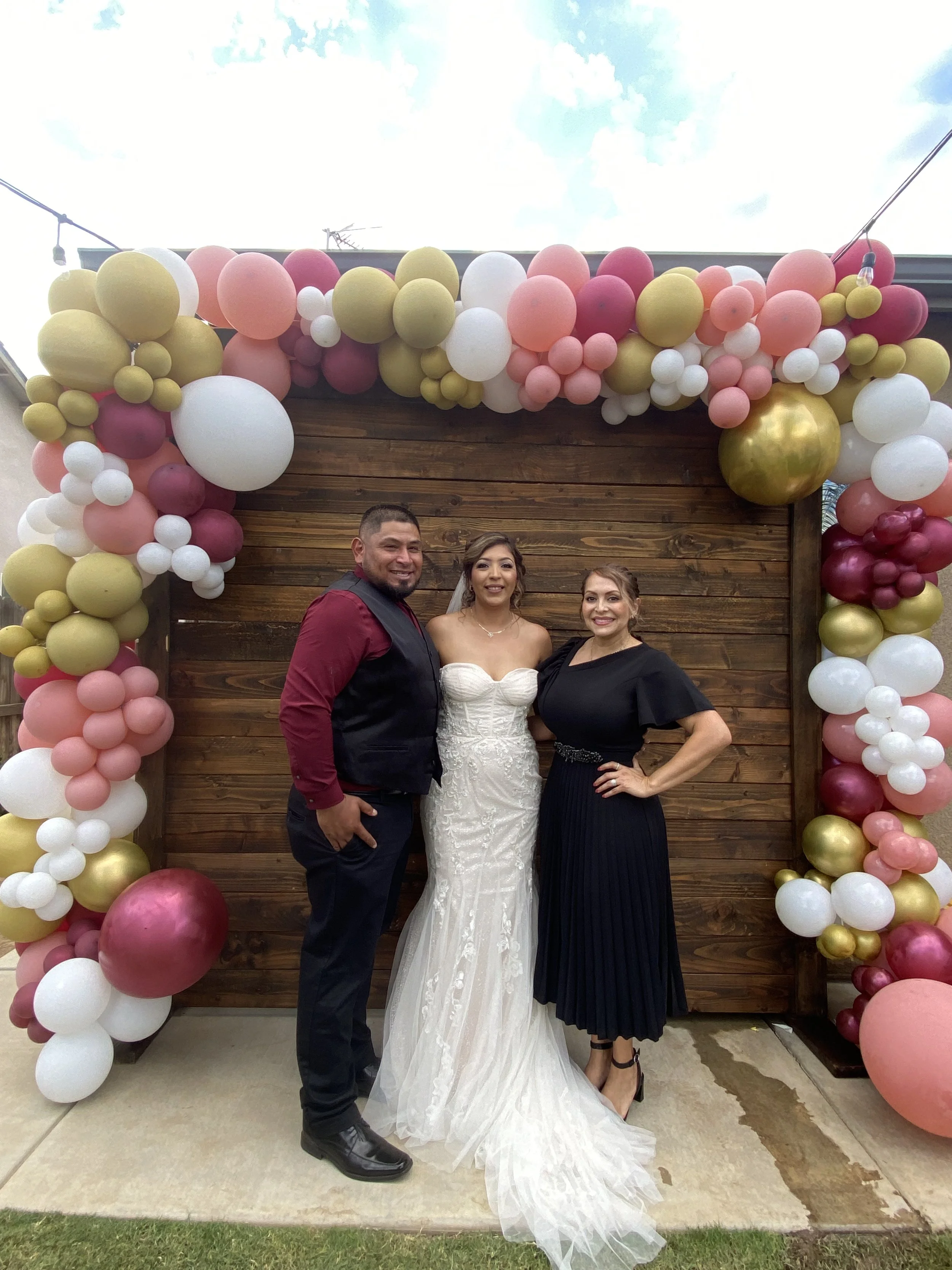Three people standing in front of a wooden backdrop adorned with pink, white, and gold balloons during a celebration.