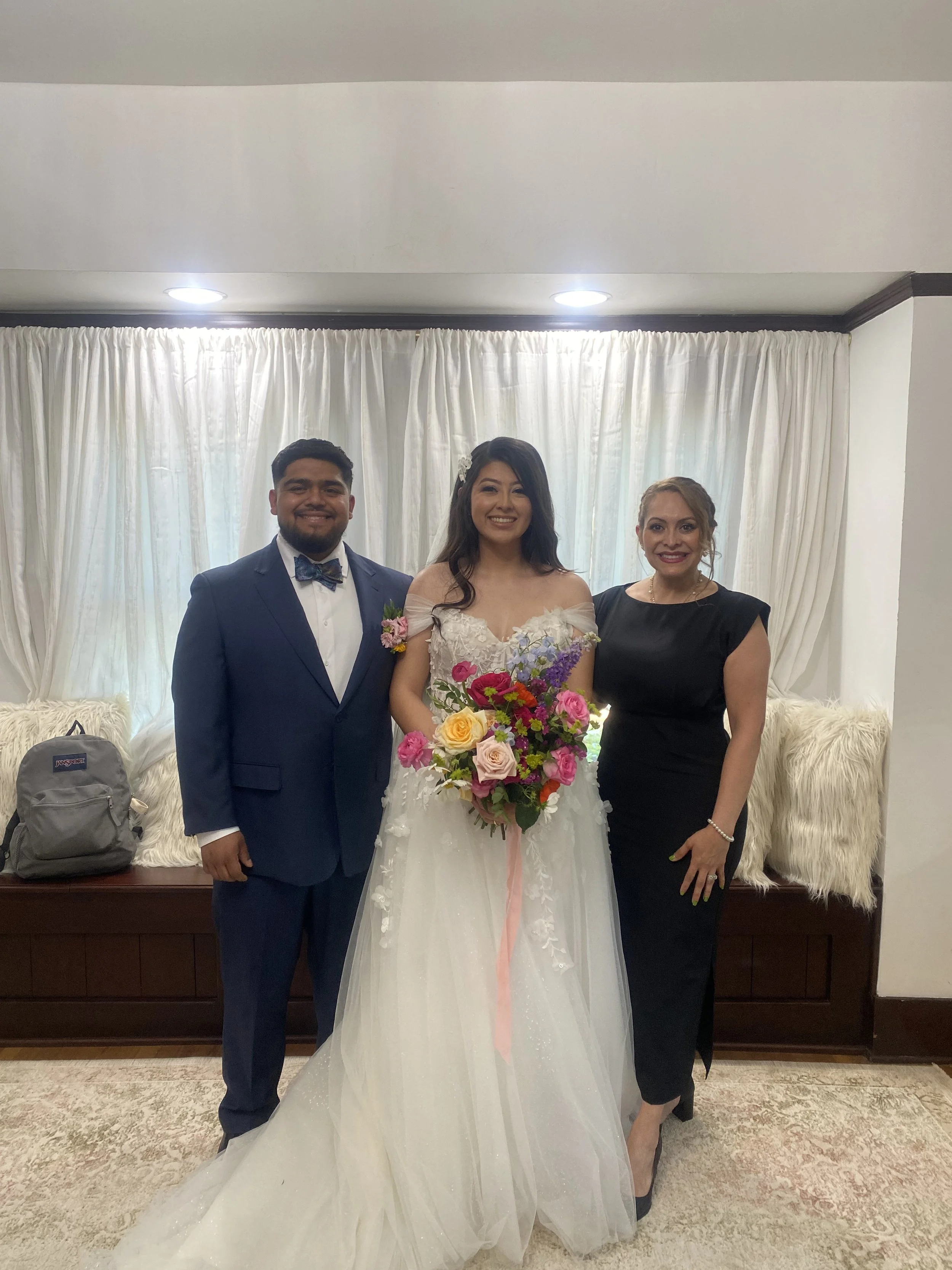 A bride in a white wedding gown holding a colorful bouquet, standing between a groom in a blue suit with a bow tie and a woman in a black dress, indoors with a white curtain backdrop.