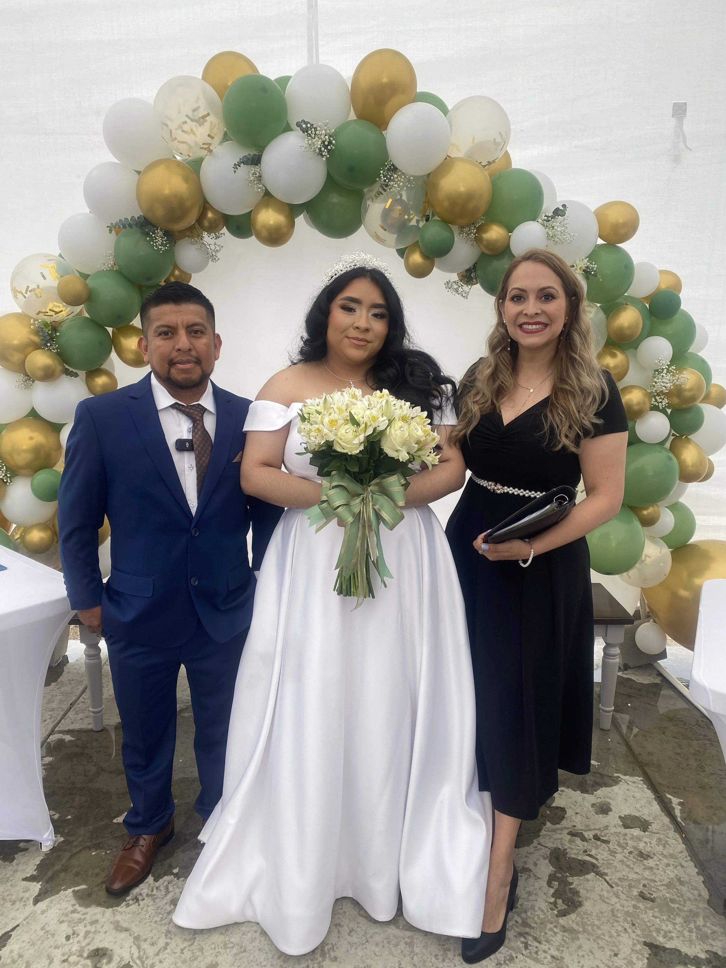 A bride in a white gown holding a bouquet of white and yellow flowers, standing between a man in a blue suit and a woman in a black dress, at a wedding event with a decorative balloon arch in green, white, and gold in the background.