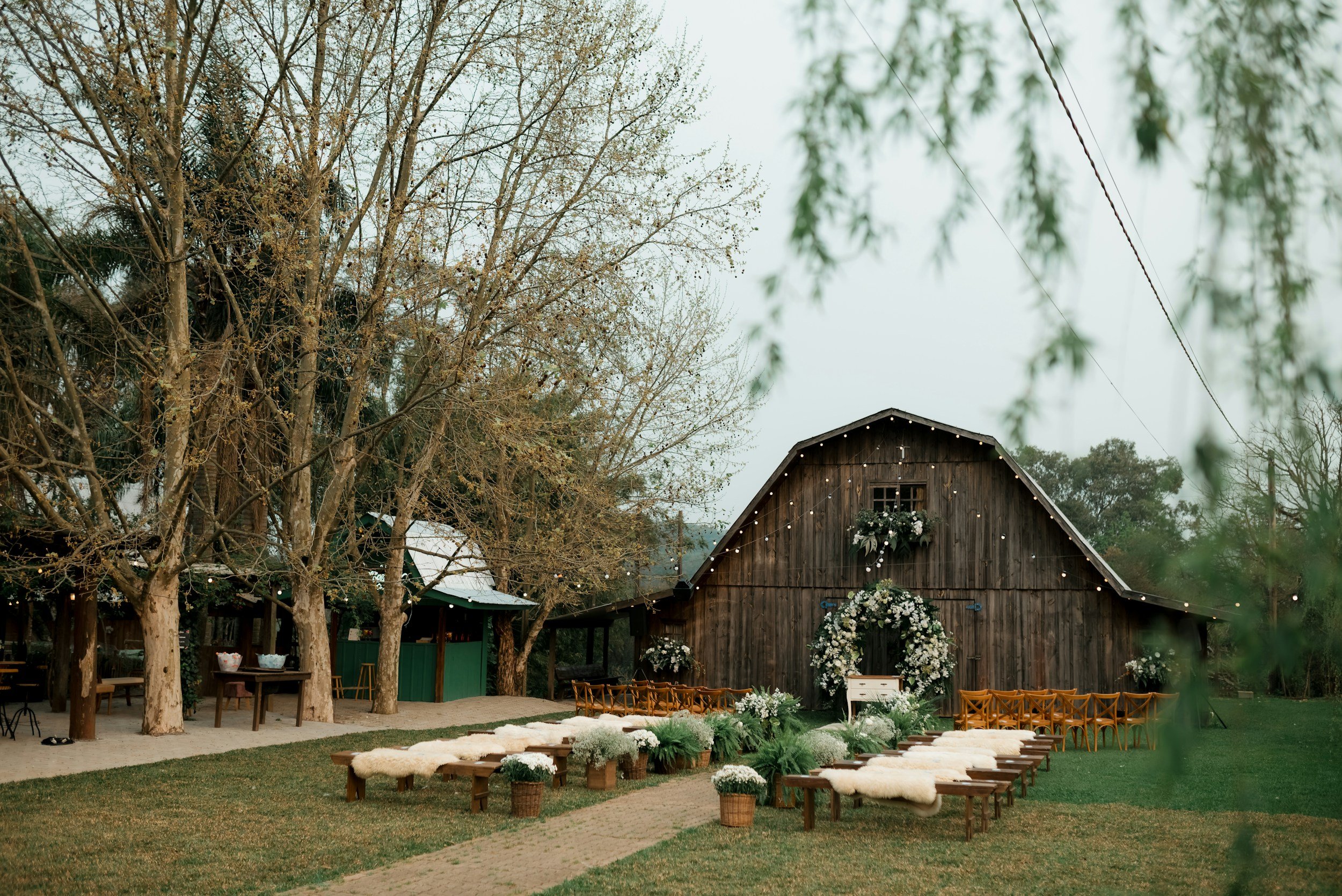 Fresno Outdoor rustic wedding setup with a wooden barn decorated with white flowers and greenery for a ceremony, surrounded by trees and arranged chairs and benches with cushions.