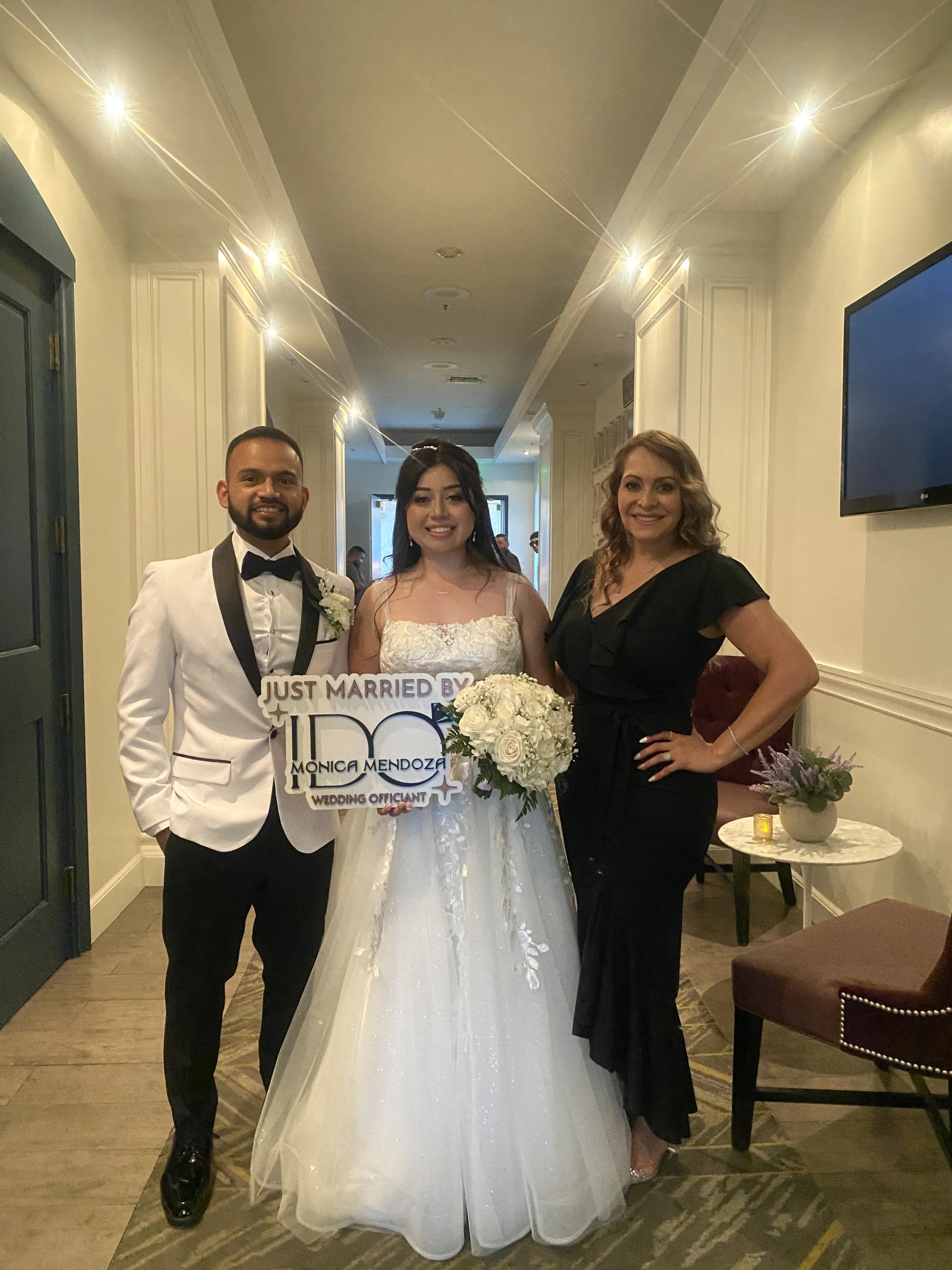 A newlywed couple and a woman standing in a hallway, smiling, with the bride holding a sign that says "Just Married by Monica Mendoza, Wedding Officiant" and a bouquet of white roses, dressed in wedding attire, with warm lighting and elegant decor.