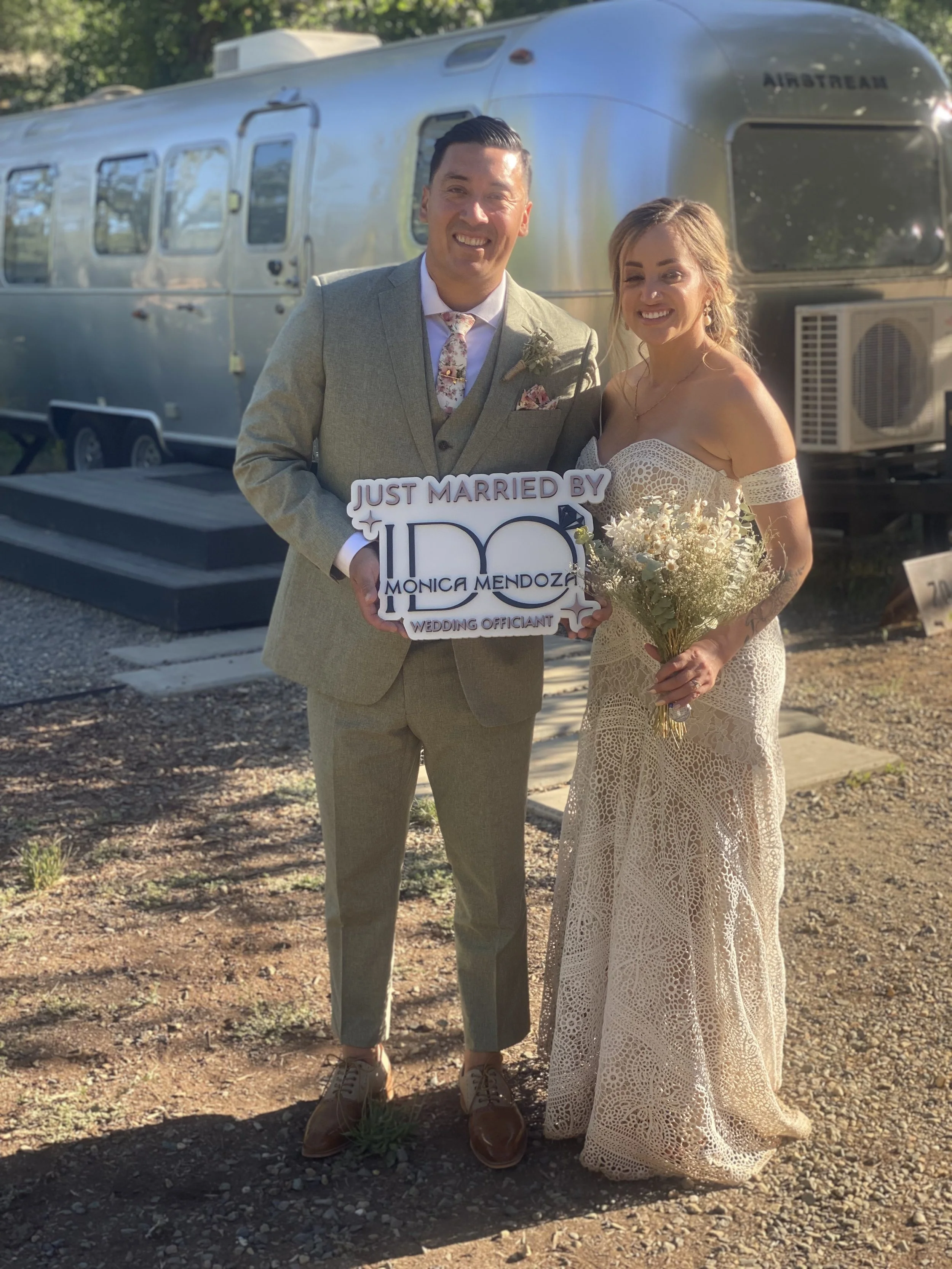 Smiling bride and groom standing outdoors in front of an Airstream trailer, holding a sign that reads "Just Married by Monica Mendoza." The groom is dressed in a light gray suit with a floral tie and pocket square, and the bride is wearing a lace off-the-shoulder wedding dress, holding a bouquet of flowers.