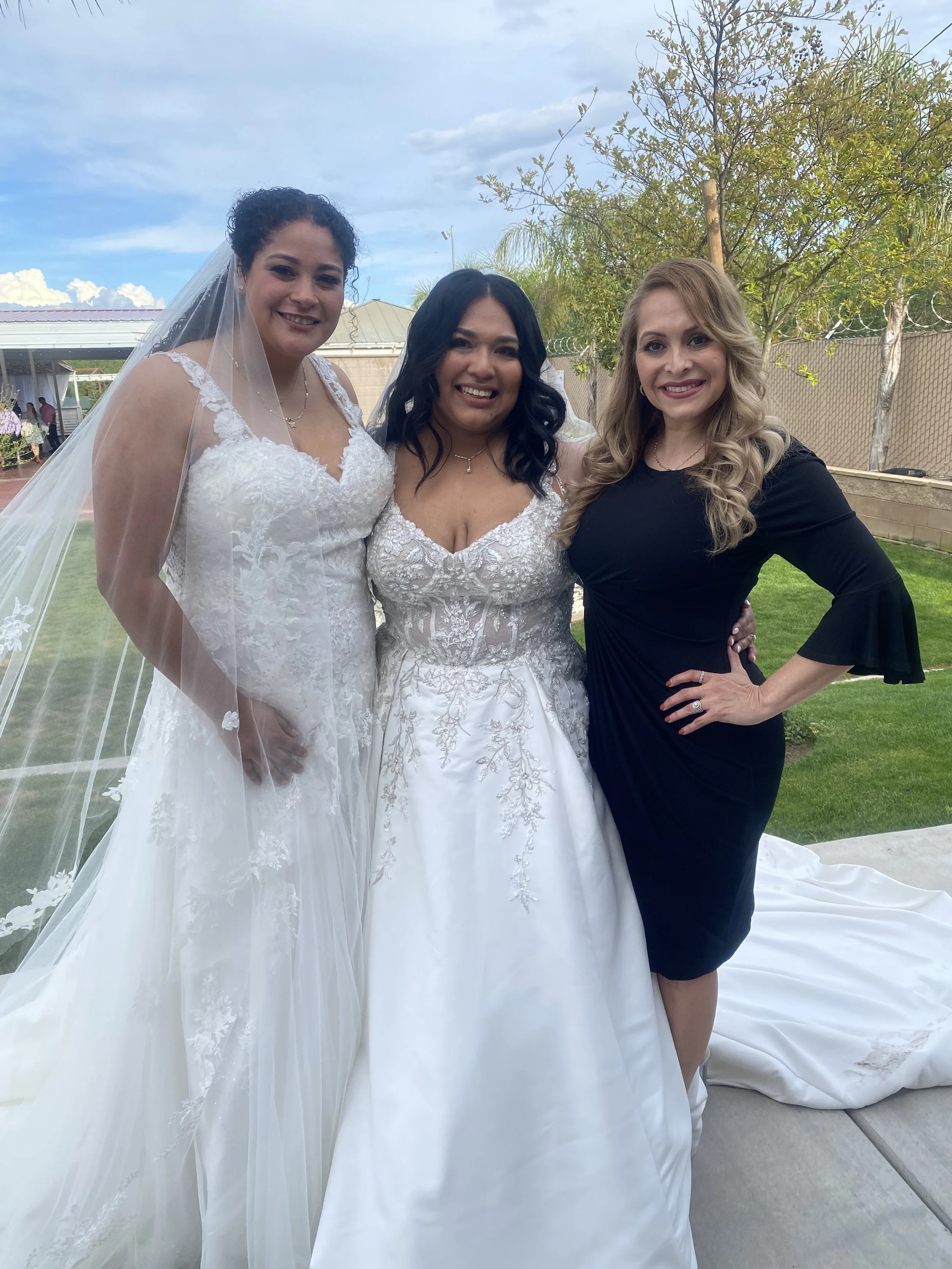 Three women standing outdoors, two in wedding dresses and one in a black dress, smiling for a photo on a cloudy day with trees and a fence in the background.