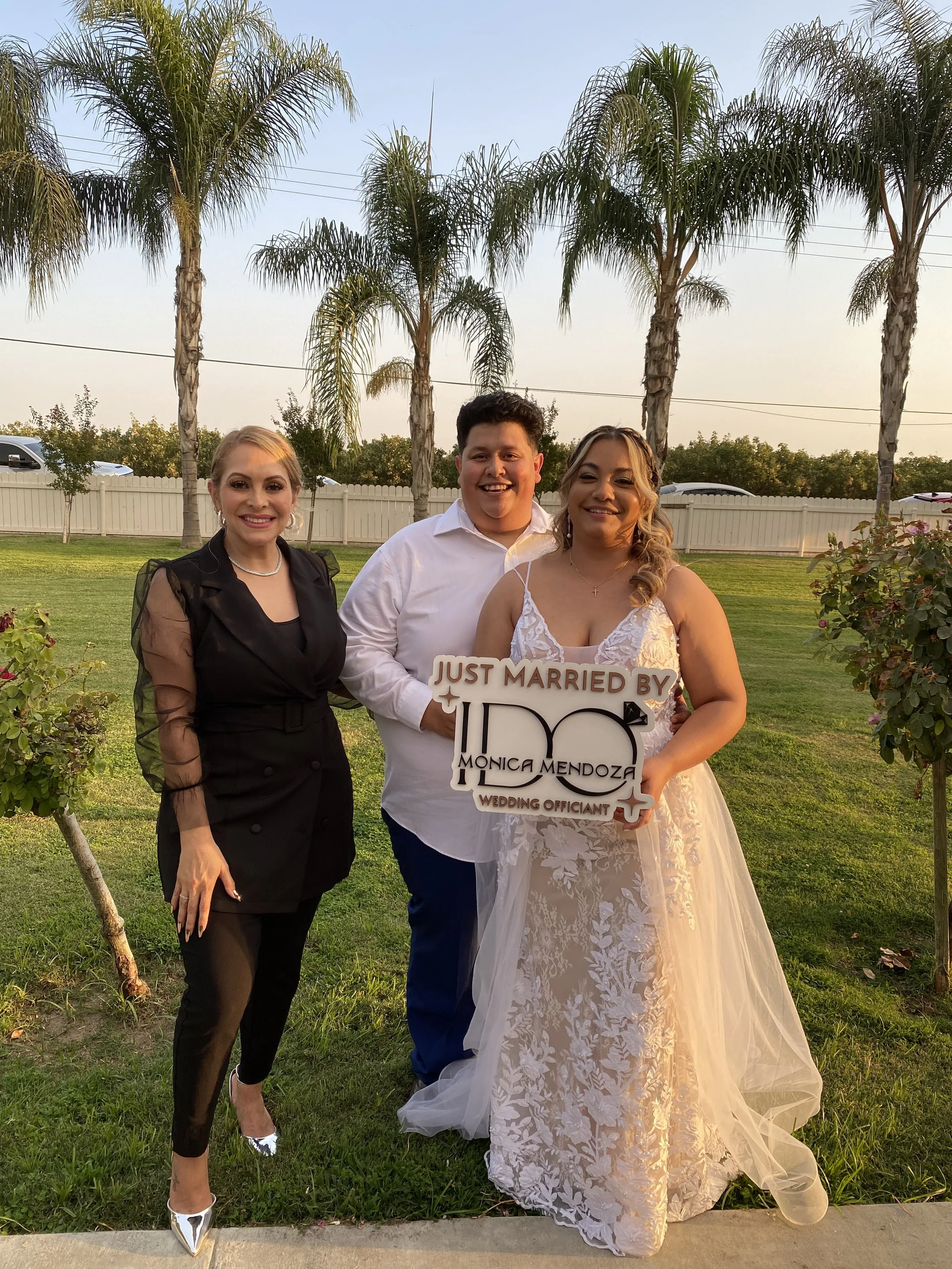 A newlywed couple smiling outdoors with a friend or family member, holding a sign that says "Just Married by Monica Mendoza Wedding Officiant." The bride is in a white lace wedding gown, and the groom and another woman are dressed in formal attire, standing on a lawn with trees and a white fence in the background.