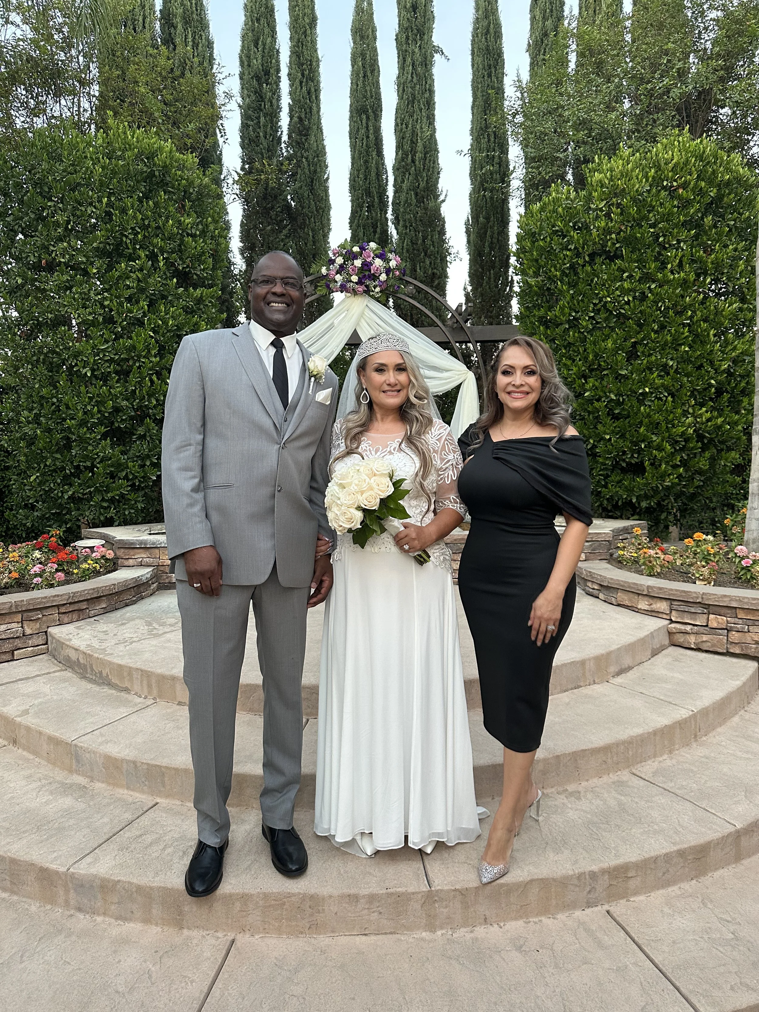 Three people standing arm-in-arm in front of a wedding altar with purple flowers and white drapery. The man on the left is wearing a gray suit, white shirt, and black tie. The woman in the center is wearing a white wedding dress and holding a bouquet of white roses. The woman on the right is dressed in a black off-the-shoulder dress and silver high heels.