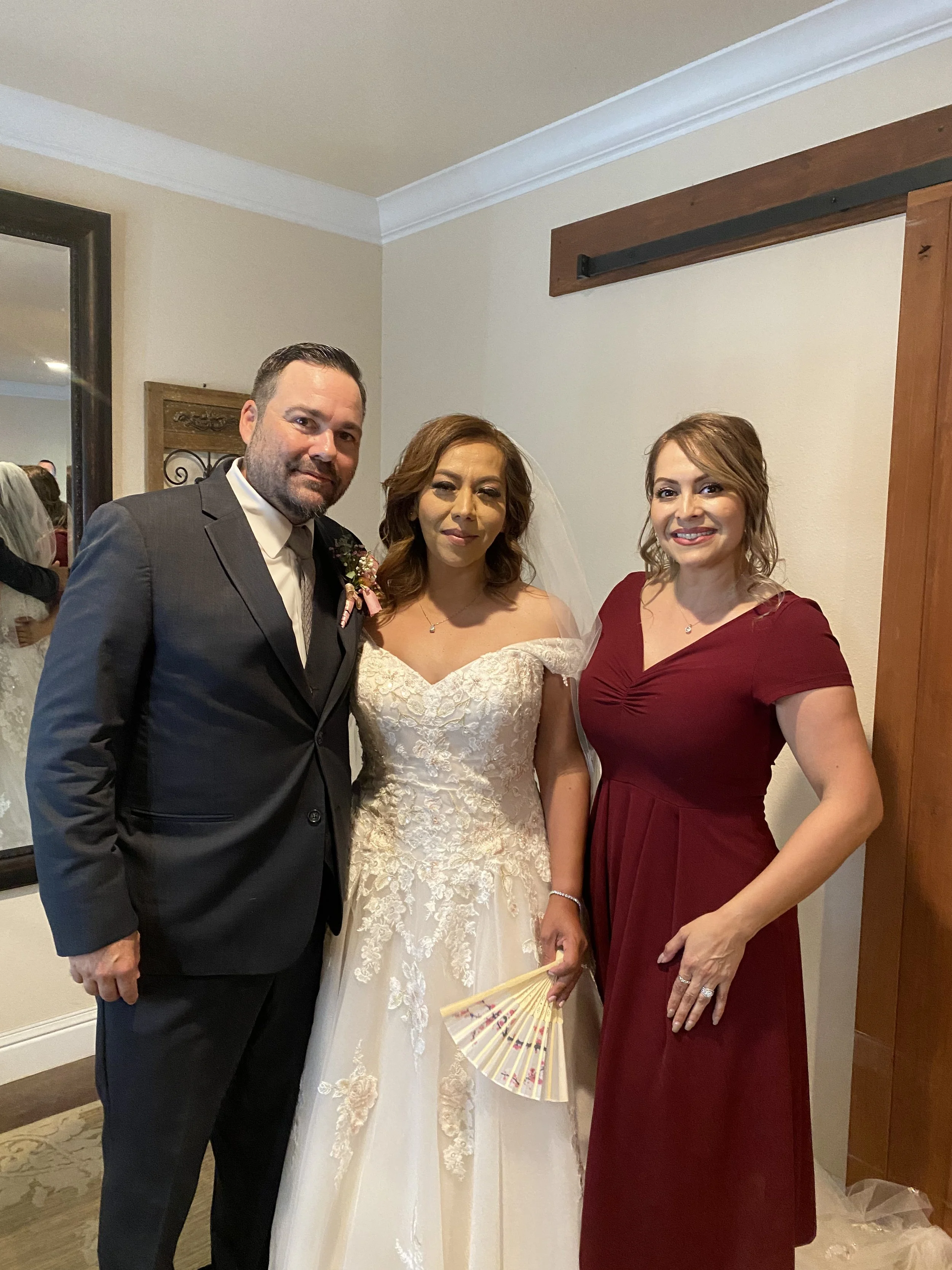 Three people dressed formally at a wedding reception, a man in a gray suit, a woman in a white wedding gown, and a woman in a red dress, standing indoors near a wall and mirror.