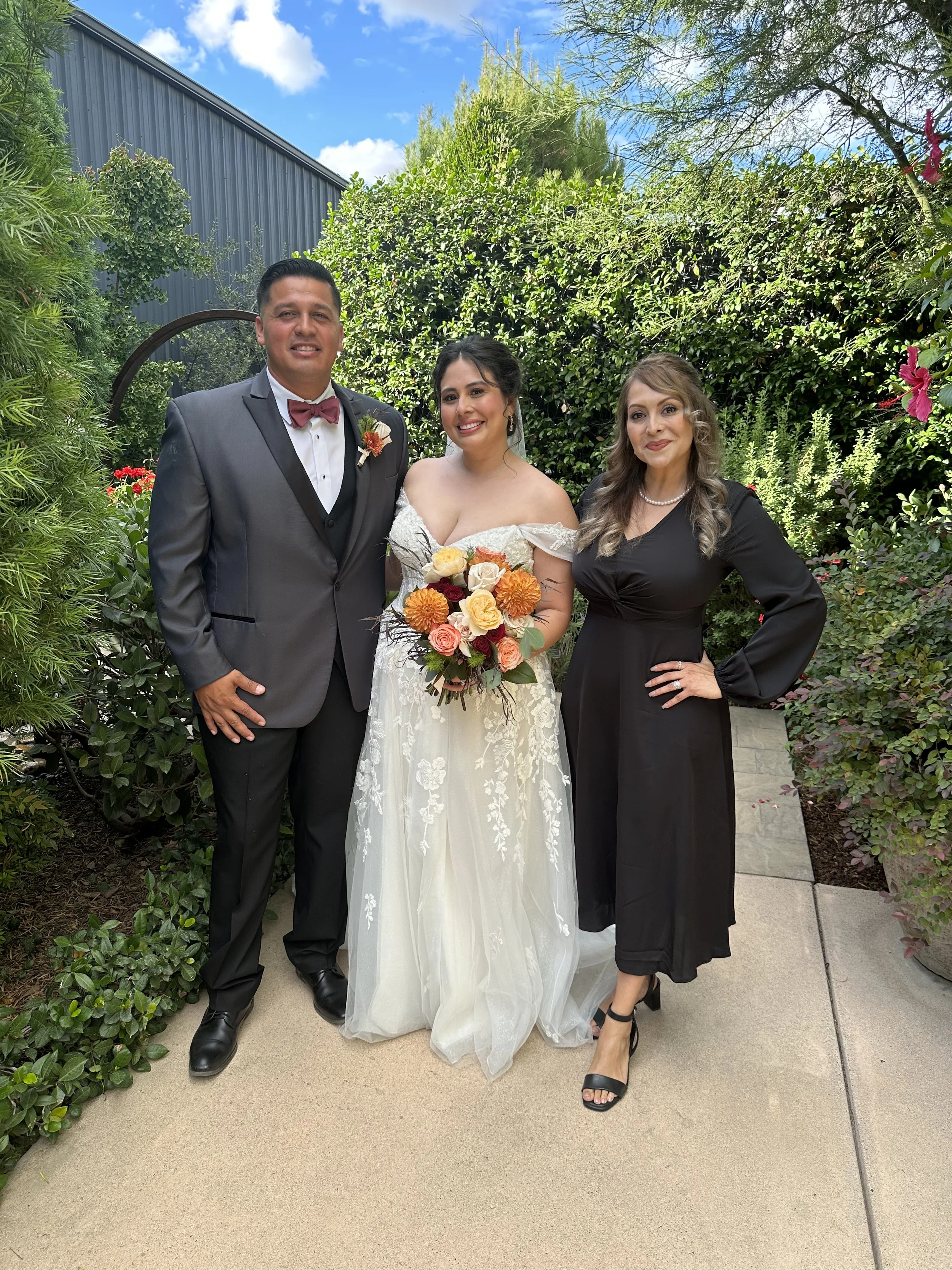 Three people in wedding attire standing on a garden path with greenery and flowers, posing for a photo during a wedding celebration.