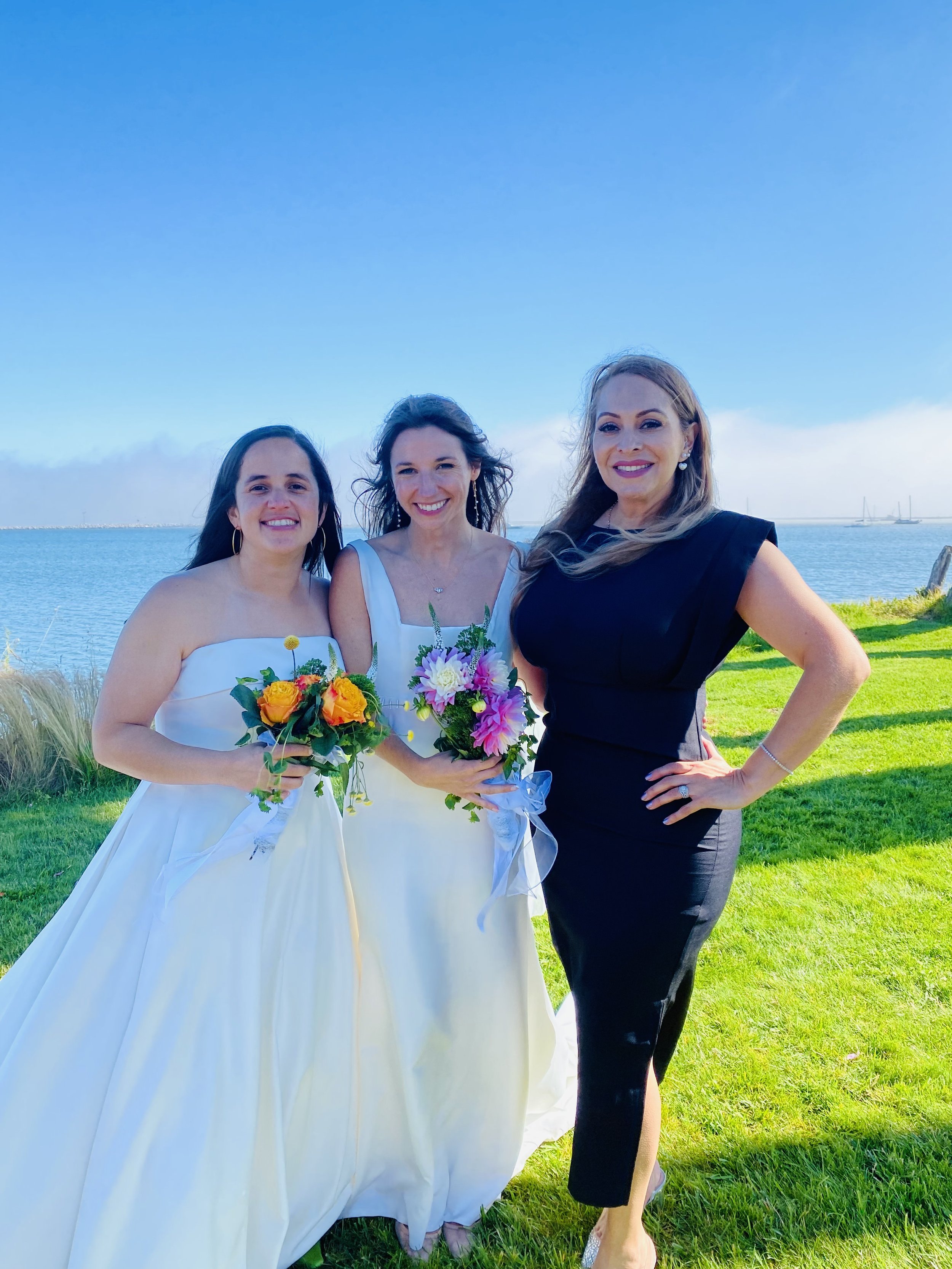 Three women by a lake, two in wedding dresses holding bouquets, and one in a black dress, smiling for the camera.