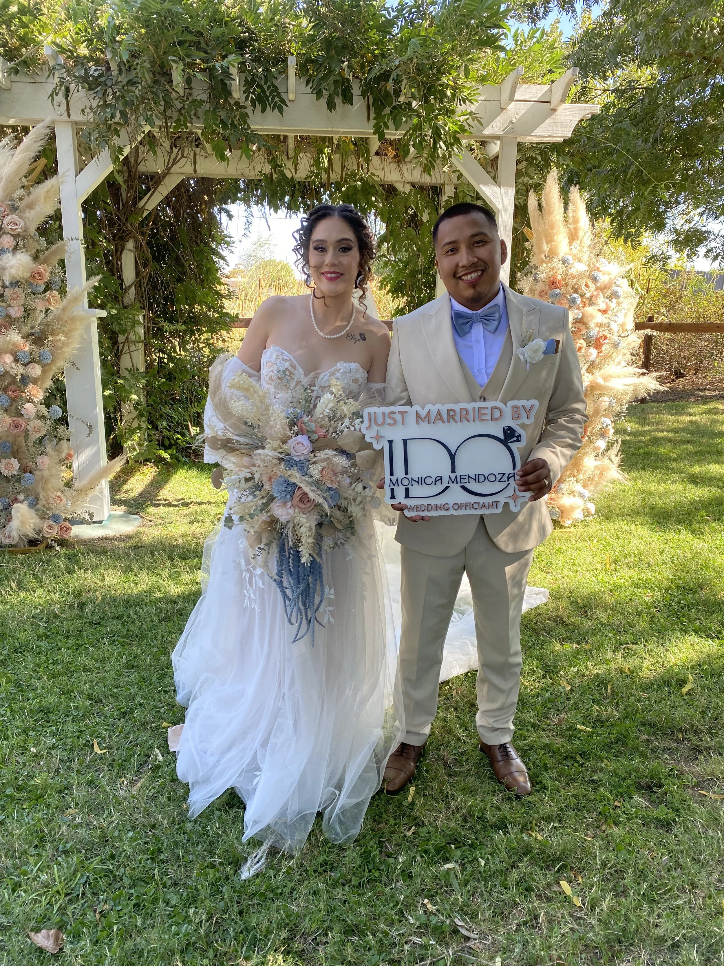 Bride and groom standing outdoors under a decorated arch, celebrating their wedding, holding a sign that says 'Just Married by Monica Mendoza, Love, WEDDING OFFICIANT'.