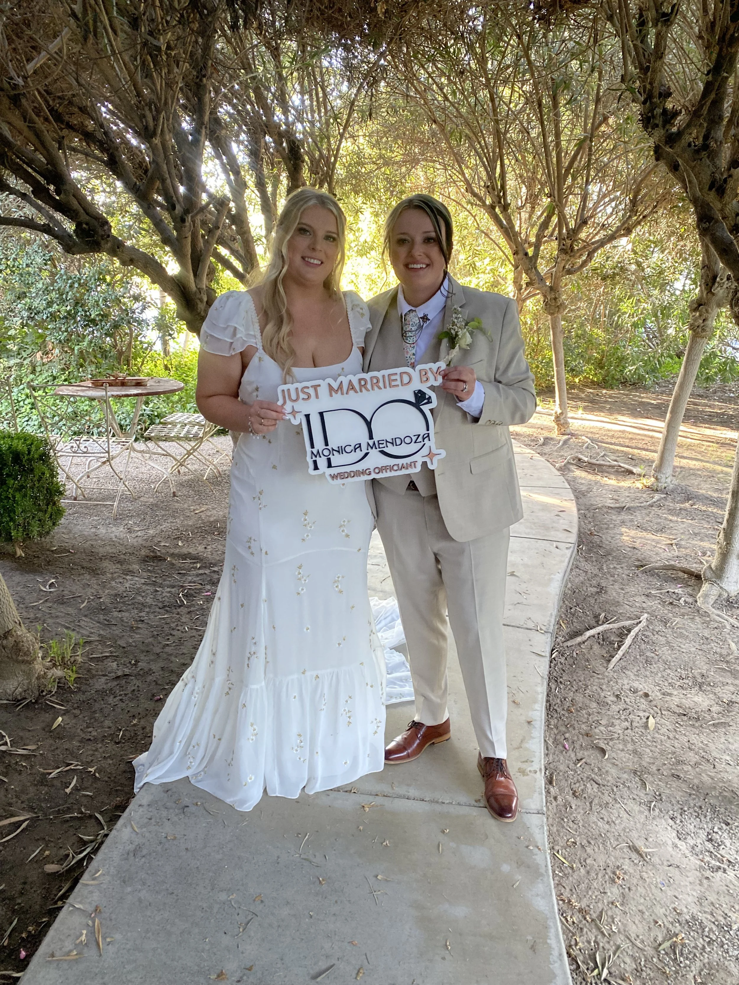 Two women standing on a sidewalk under trees, holding a sign that reads "Just Married" with the names Monica Mendoza and a wedding officiant, smiling at the camera.