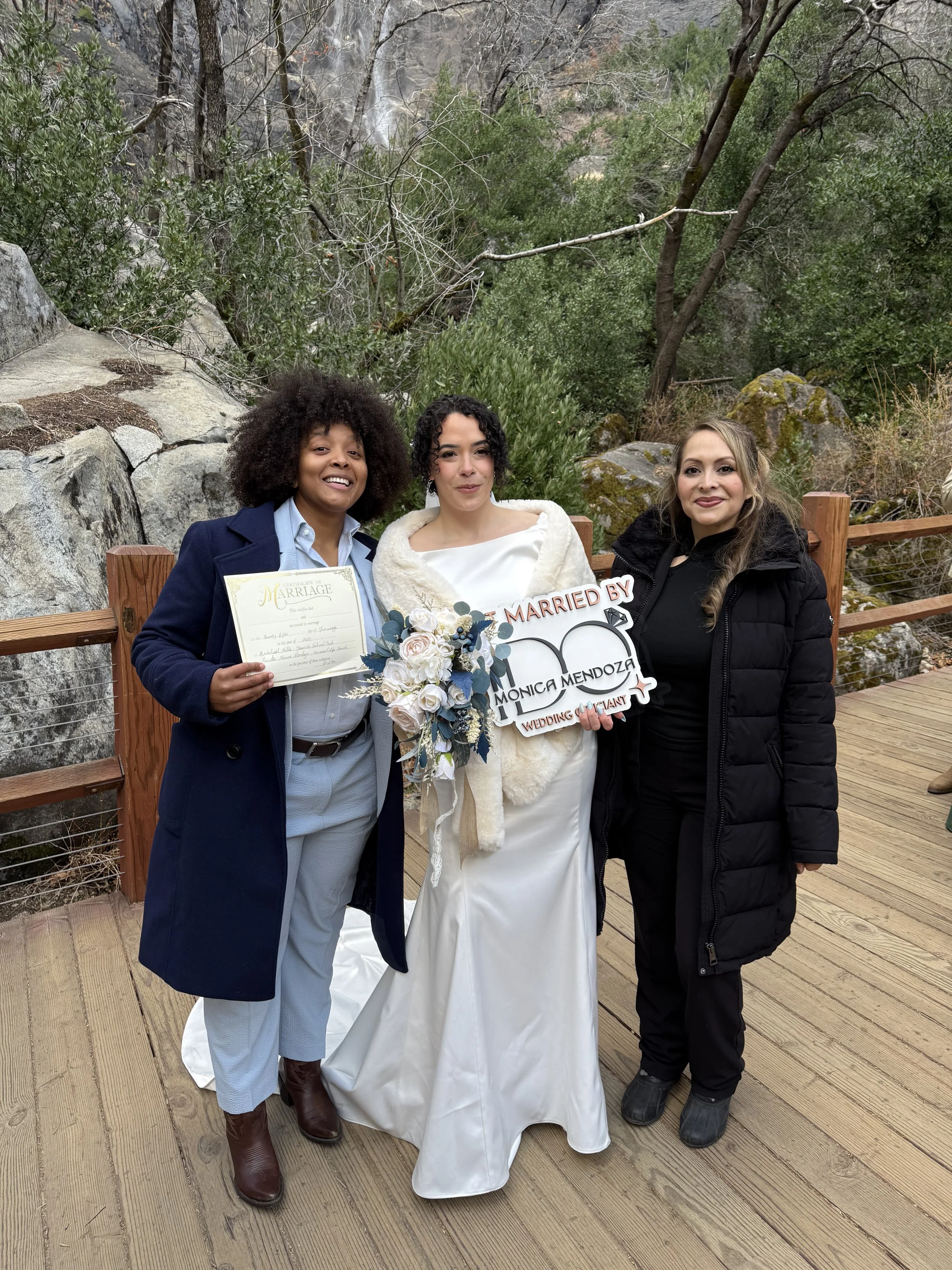 Three women standing outdoors on a wooden platform, celebrating a wedding. The woman in the middle is dressed in a white wedding gown holding a bouquet, and the other two women are wearing coats. The woman on the left is holding a marriage certificate, and the woman on the right is holding a sign that reads, 'MARRIED BY MONICA MENDEZ, WEDDING OFFICIANT.' They are smiling and posing for the photo, with a natural background of rocks and trees.