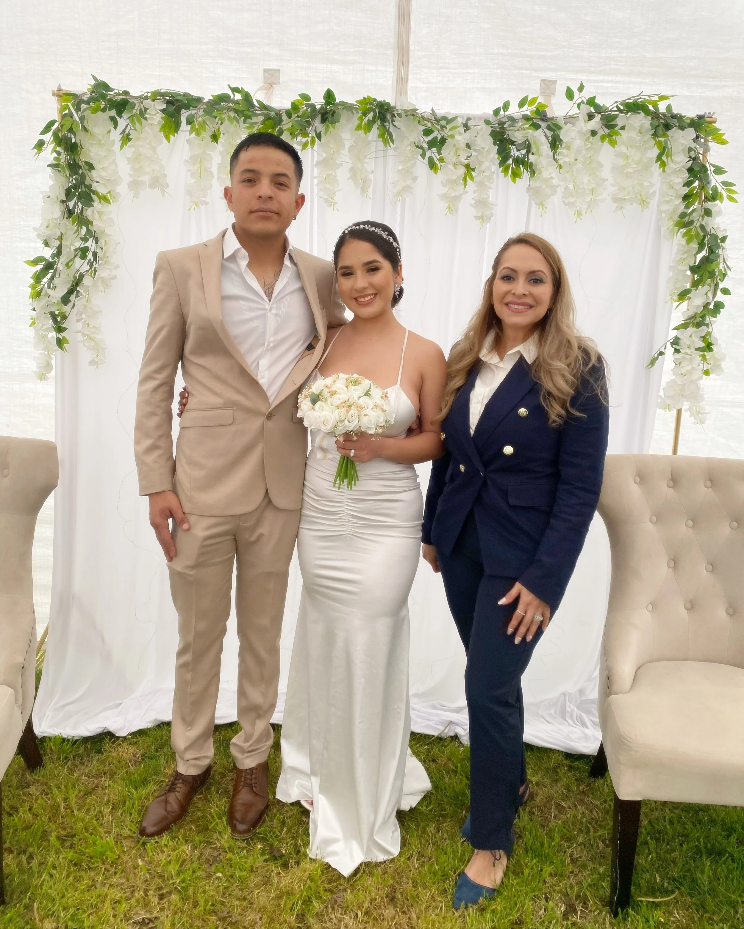 A wedding couple with a woman, standing outdoors in front of a white backdrop decorated with greenery and white flowers, with two beige chairs on each side, on grass.