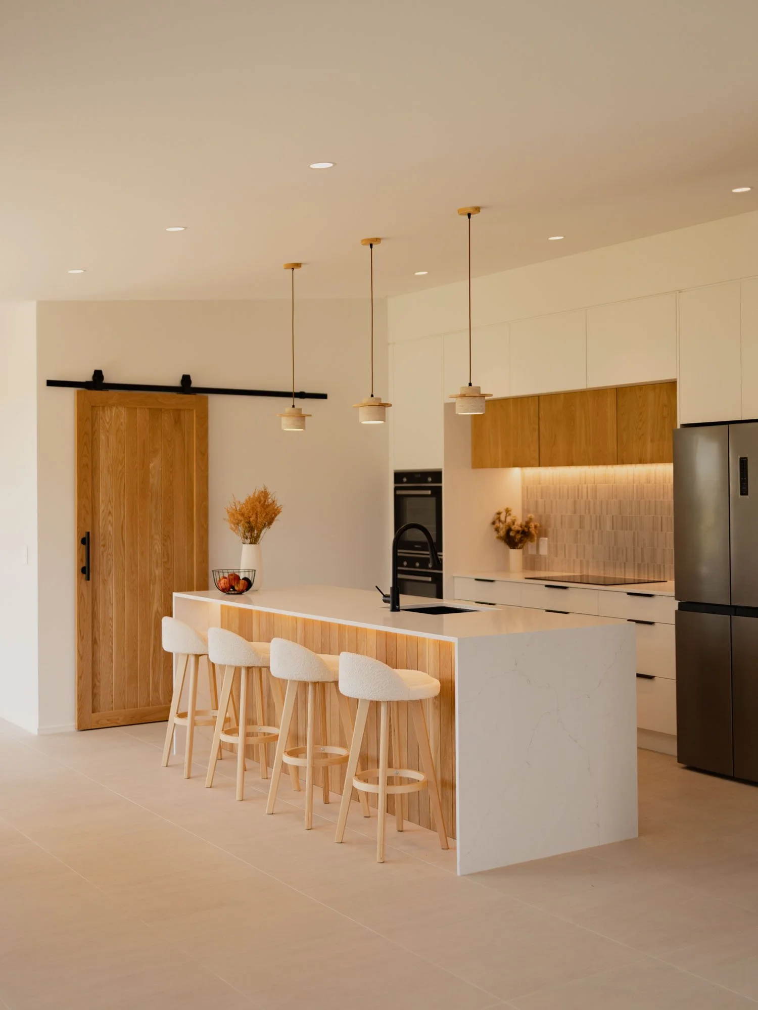 Modern kitchen with white countertops, wooden cabinets, and a black sink. Three pendant lights hang above the island, which has four white bar stools. There is a sliding barn door and some decorative plants by Ayline Builders - Taupō NZ.