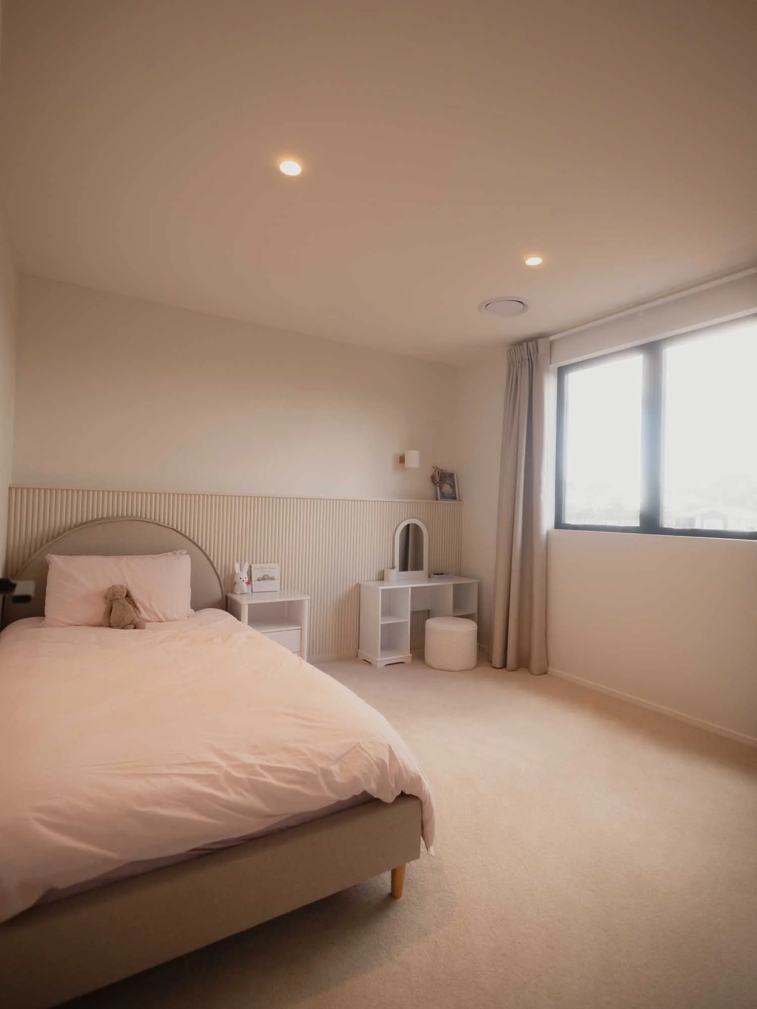 A cozy bedroom with a bed, side table with stuffed animal, vanity table with mirror, small stool, large window with curtains, ceiling lights, and cream-colored walls and carpet, with custom panelling by Ayline Builders - Taupō NZ.