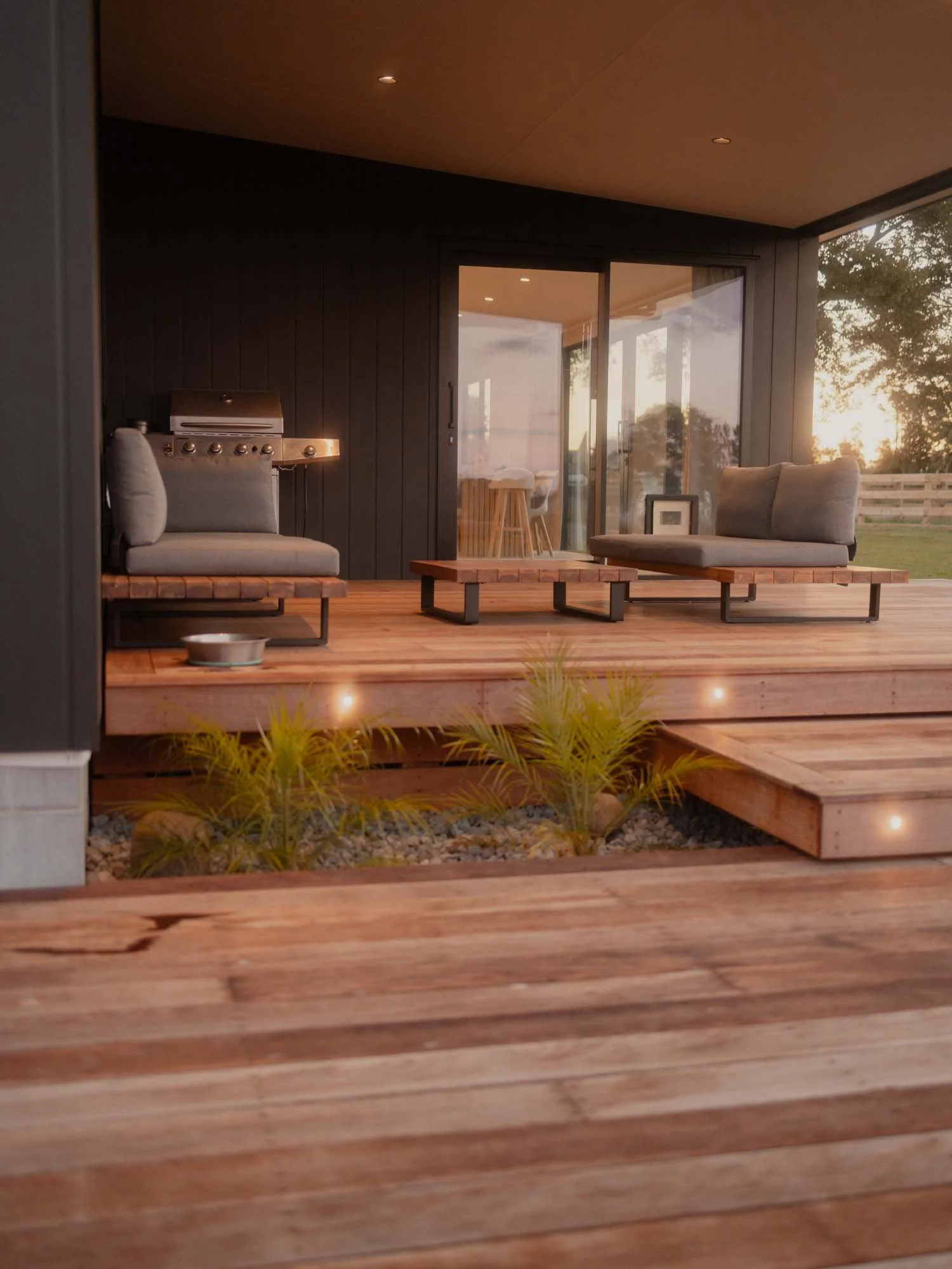 Outdoor wooden deck with two modern cushioned chairs, a small table, a barbecue grill, and a sliding glass door leading to an interior area with barstools at a counter, during sunset by Ayline Builders - Taupō NZ.
