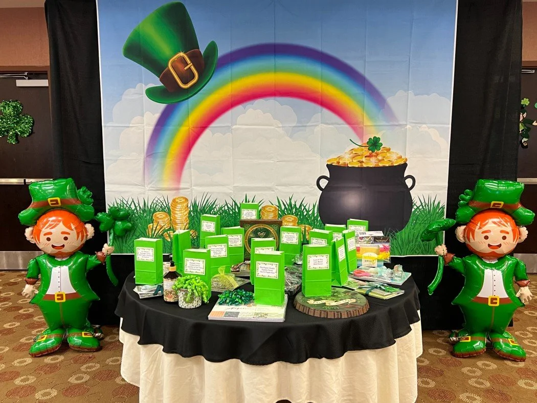 St. Patrick's Day celebration display with two balloon leprechauns, a rainbow, a pot of gold, and various small green-themed items on a round table.
