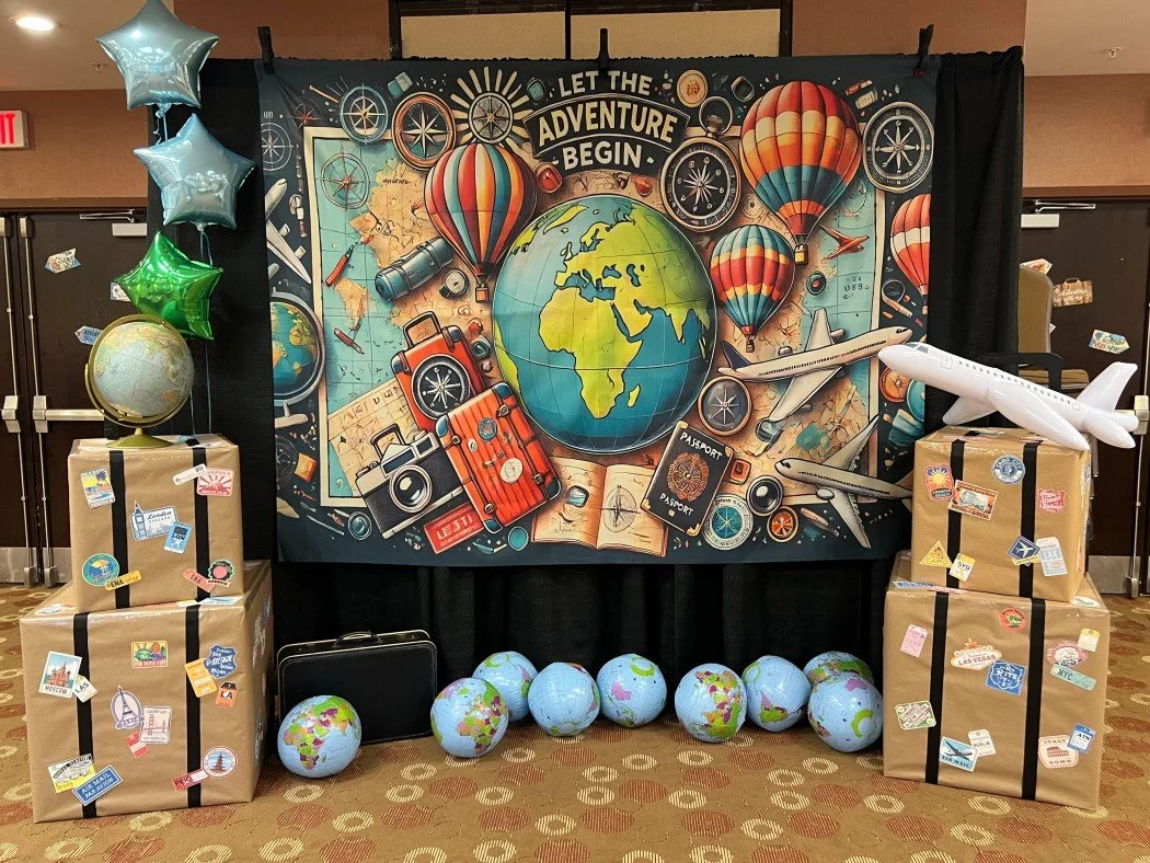 Travel-themed display with a large backdrop featuring a globe, hot air balloons, airplanes, a camera, a suitcase, a map, and travel essentials with the banner text "Let the adventure begin." In front, there are globe balloons, cardboard boxes with tr