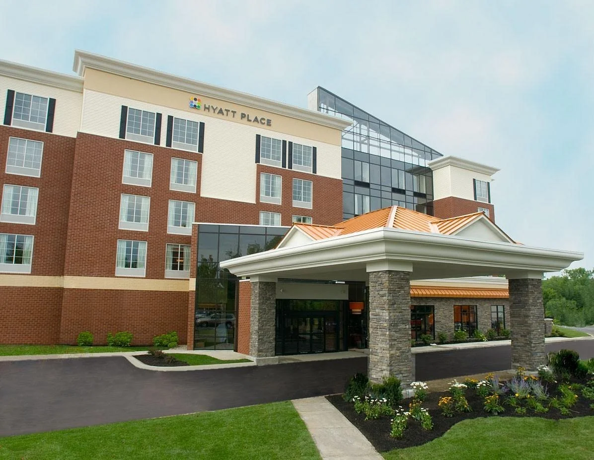 Exterior of Hyatt Place hotel with brick and glass facade, landscaped front, and covered entrance with stone pillars.