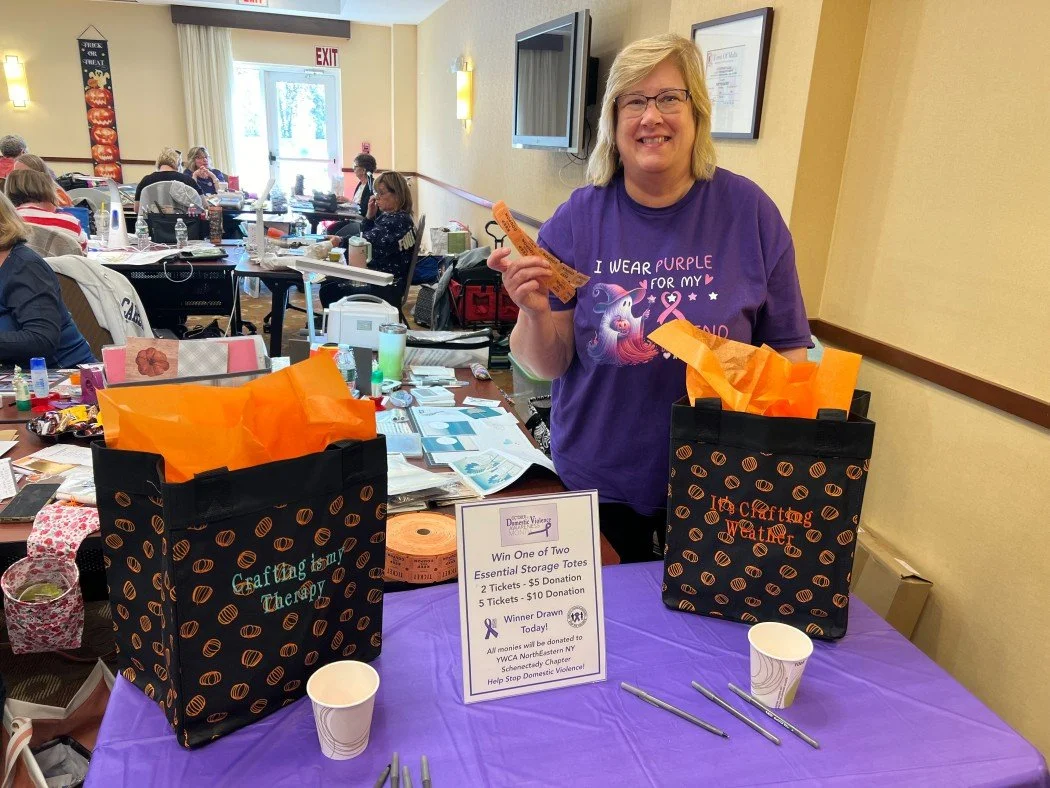 A woman in a purple shirt smiling at a table with gift bags, donation sign, and raffle tickets in a decorated event room filled with people.