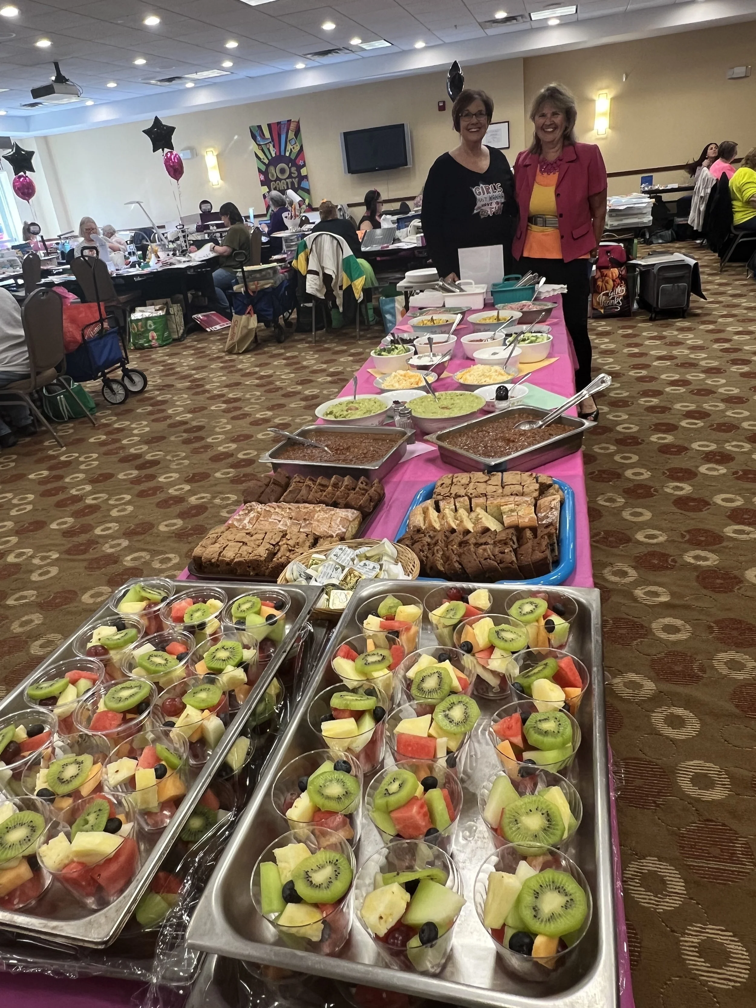 Food buffet table with fruit cups, cakes, and other dishes at an indoor event with women standing nearby