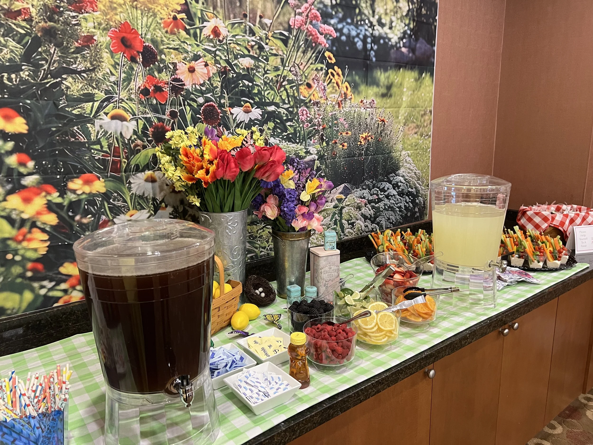 A refreshment table with colorful flowers, fresh fruits, and beverages in large dispensers, set against a bright floral mural.