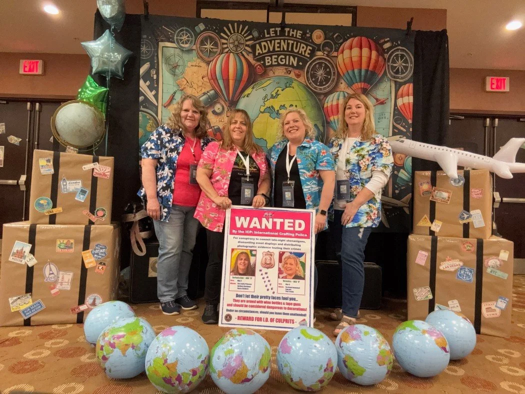 Four women in Hawaiian shirts standing behind a large pink 'Wanted' poster with pictures of two women, surrounded by globe decorations, suitcases, balloons, and an airplane model, with a travel-themed backdrop.
