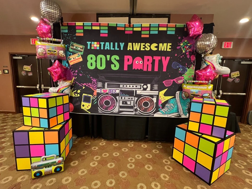 Decorative 80's themed party setup with a colorful backdrop reading "Totally Awesome 80's Party" featuring images of boomboxes, cassette tapes, a handheld gaming console, sunglasses, a sneaker, and balloons shaped like hearts and disco balls, with tw