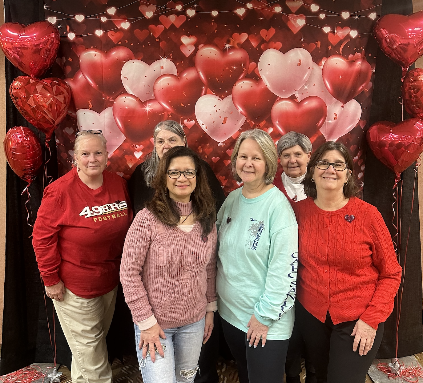 Six women standing in front of a Valentine's Day themed backdrop with heart-shaped balloons and decorations, smiling at the camera.