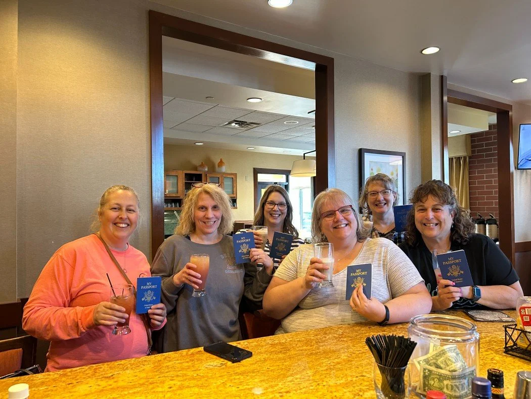 Seven women sitting at a restaurant table, smiling and holding drinks and passports, celebrating together.
