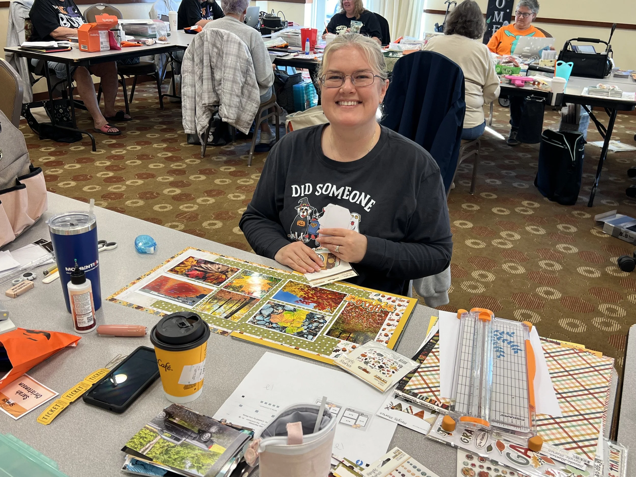 A woman sitting at a table in a craft room, smiling, with various scrapbook supplies, photos, and decorations on the table, and other people working on crafts in the background.