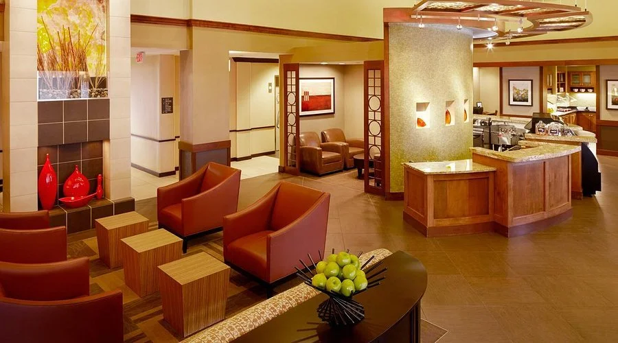 Hotel lobby with leather armchairs, wooden side tables, decorative vases, a check-in counter, and a seating area with artwork on the walls.