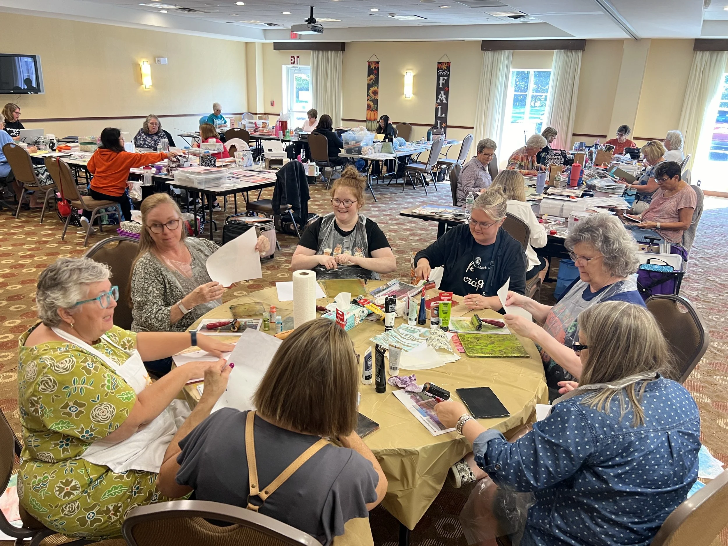 Group of women participating in a crafting workshop around a table in a well-lit room, with various supplies and decorations indicating a community or recreational event.
