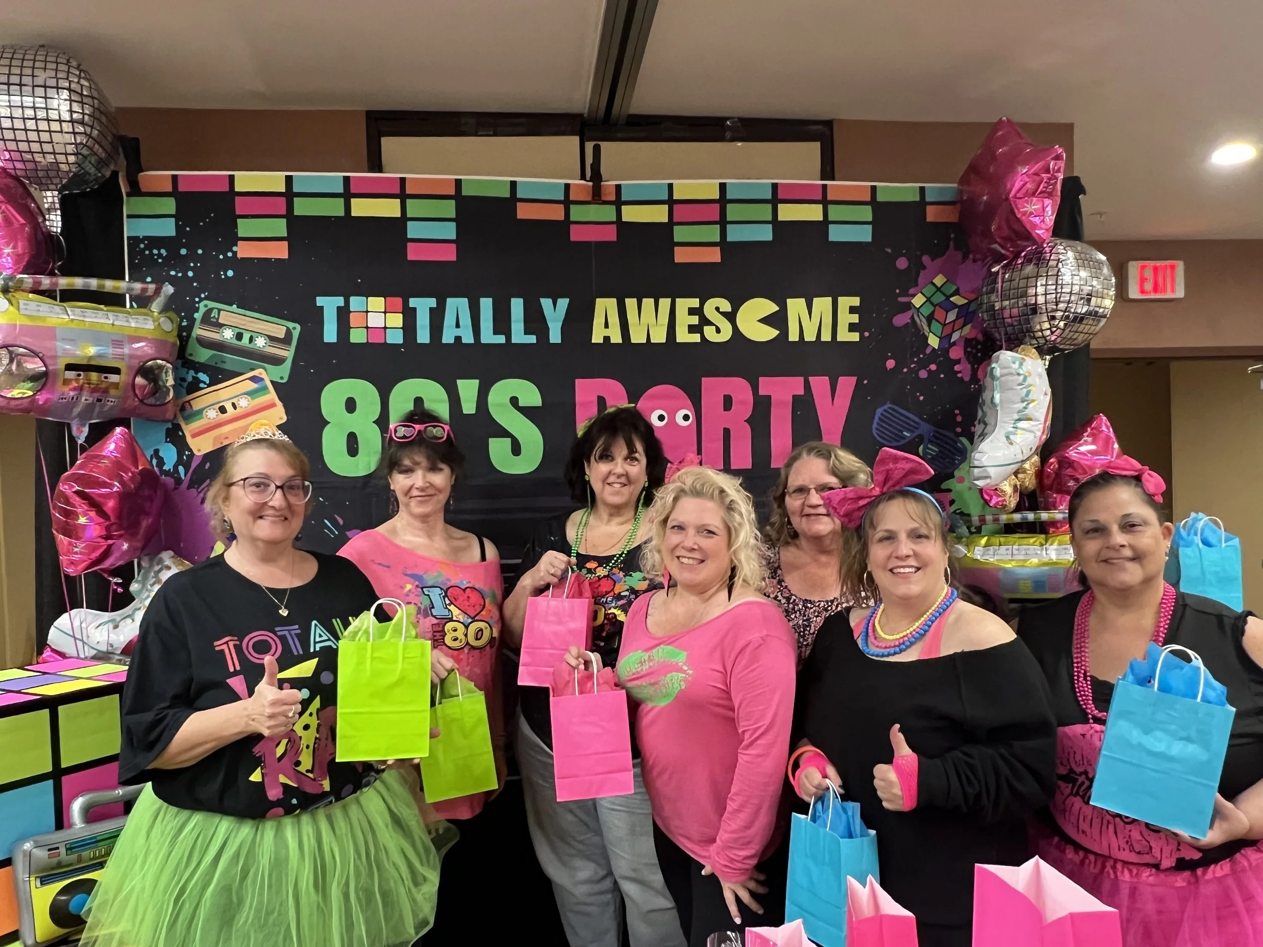 Seven women smiling and posing for a photo at an 80s themed party in front of a colorful backdrop with retro cassette tapes, sunglasses, and boombox graphics. They are dressed in 80s-inspired clothing and accessories, holding gift bags.