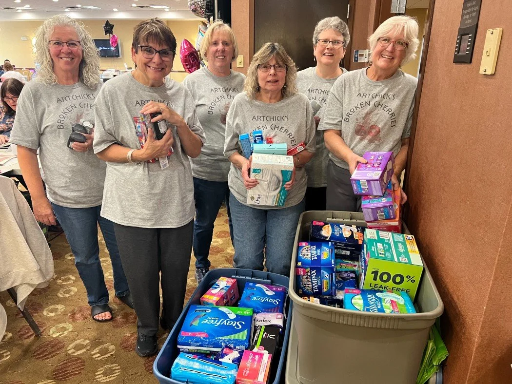 Six women stand behind bins filled with hygiene products at a donation event, all smiling and wearing matching gray T-shirts with a print referencing cherry flavor.