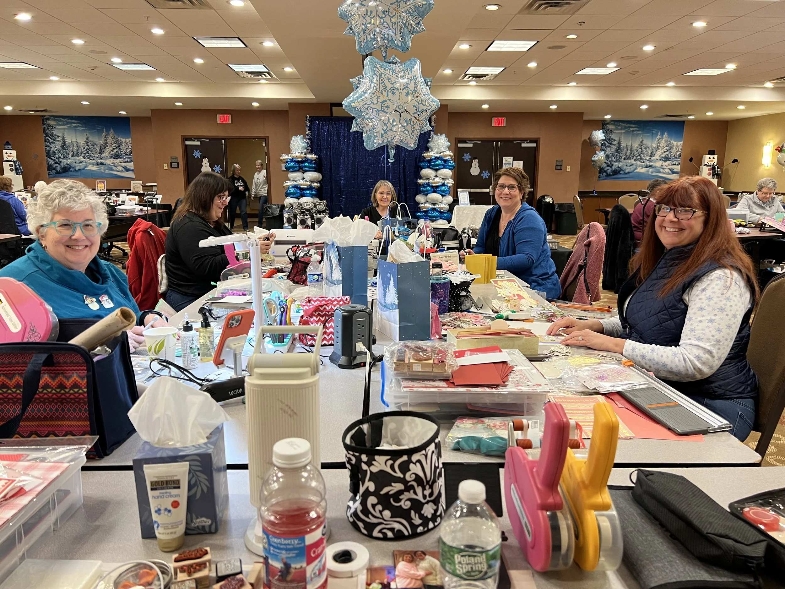 Four women sitting at a long table decorated with various craft supplies, holiday decorations, and gifts, in a festive, decorated room with snowflake ornaments and a winter scene backdrop.