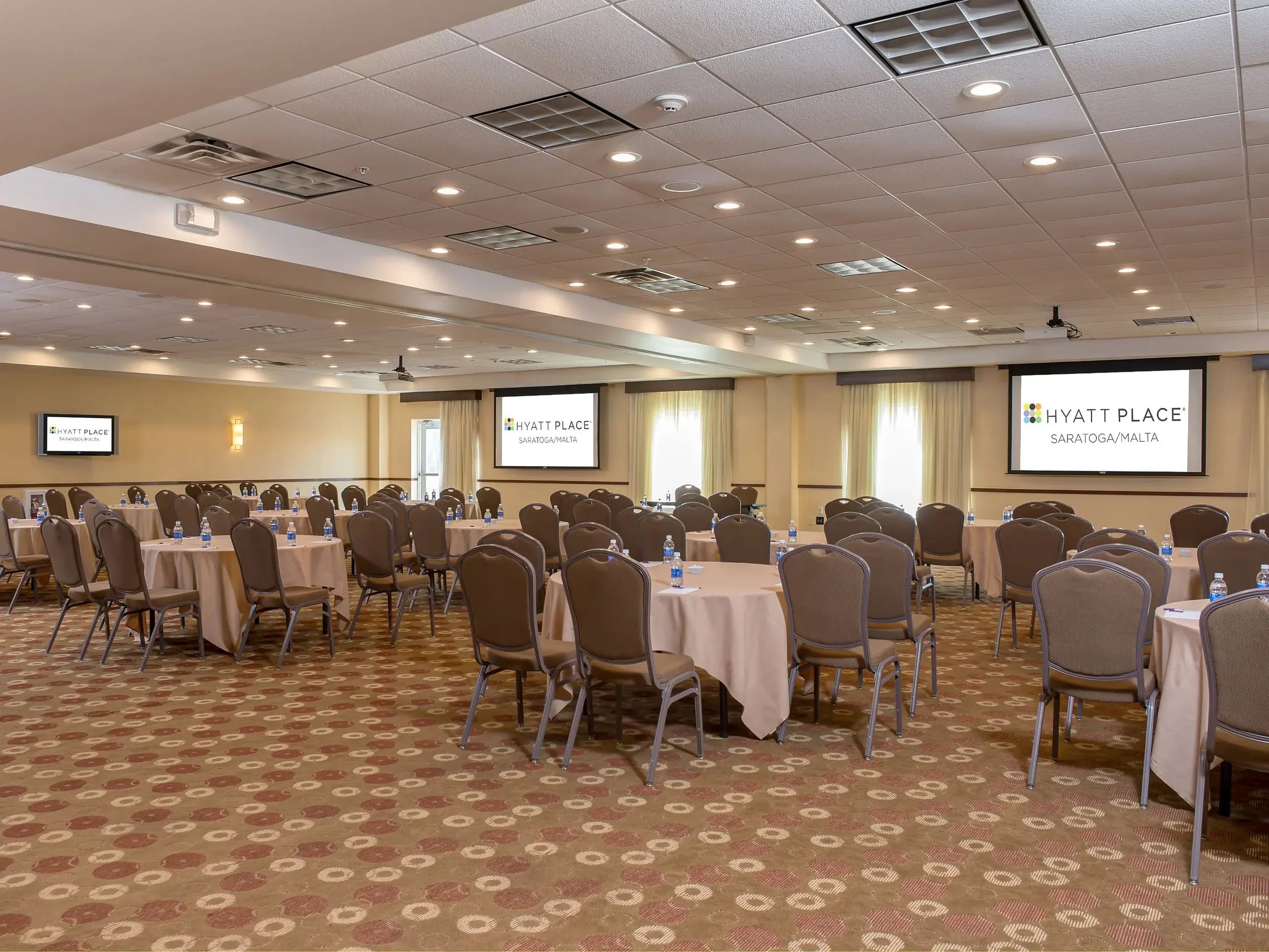 Conference room set up with round tables covered in white tablecloths, chairs around each table, and water bottles at each seat. There are three large screens displaying the Hyatt Place logo and name, with the location Saratoga/Malta. The room has be