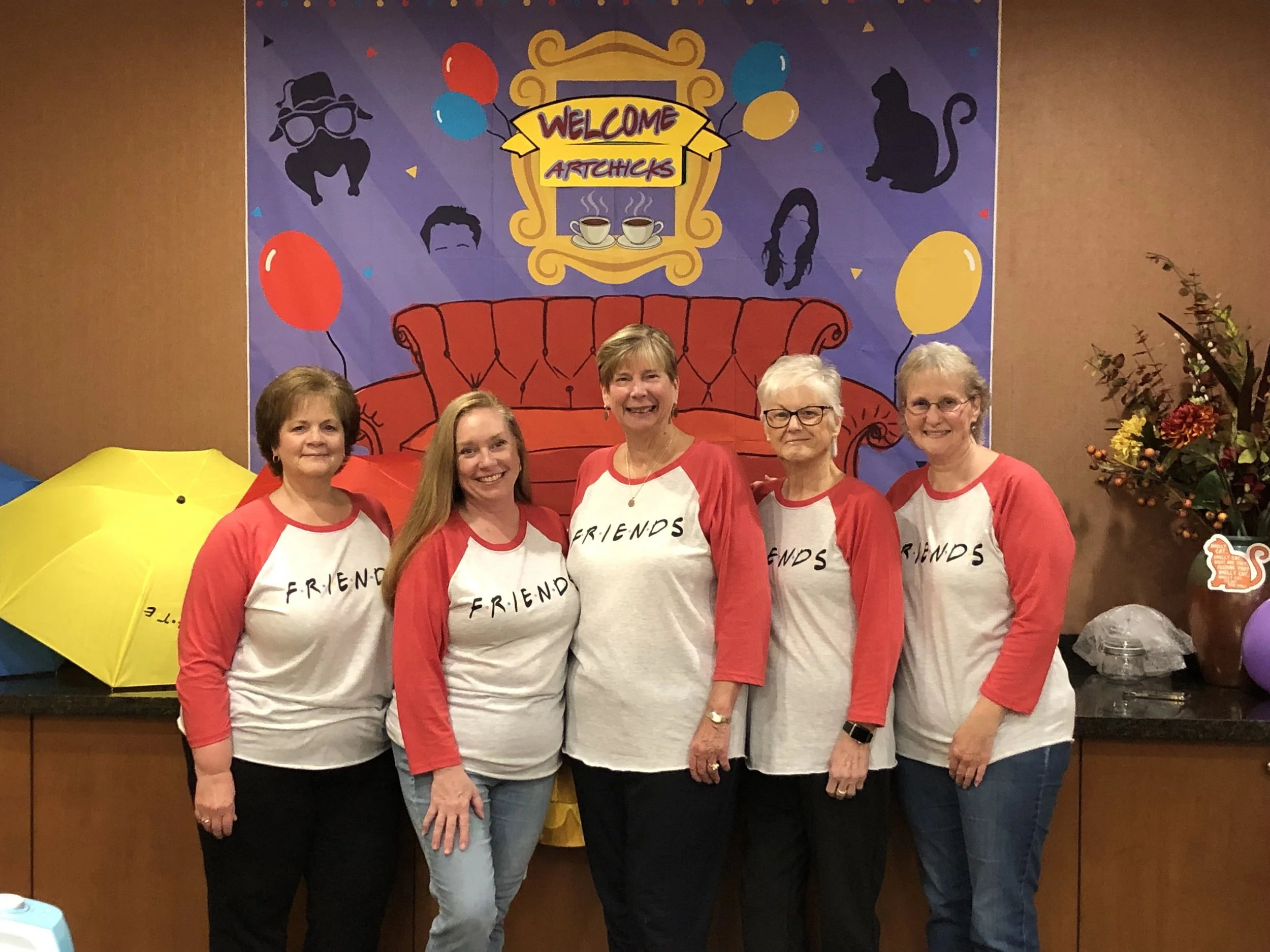 Five women are standing side by side in front of a colorful festive backdrop with balloons, a sofa illustration, and a sign that says 'Welcome Argitches.' They are all wearing matching white and red shirts with the word 'FRIENDS' printed on the front