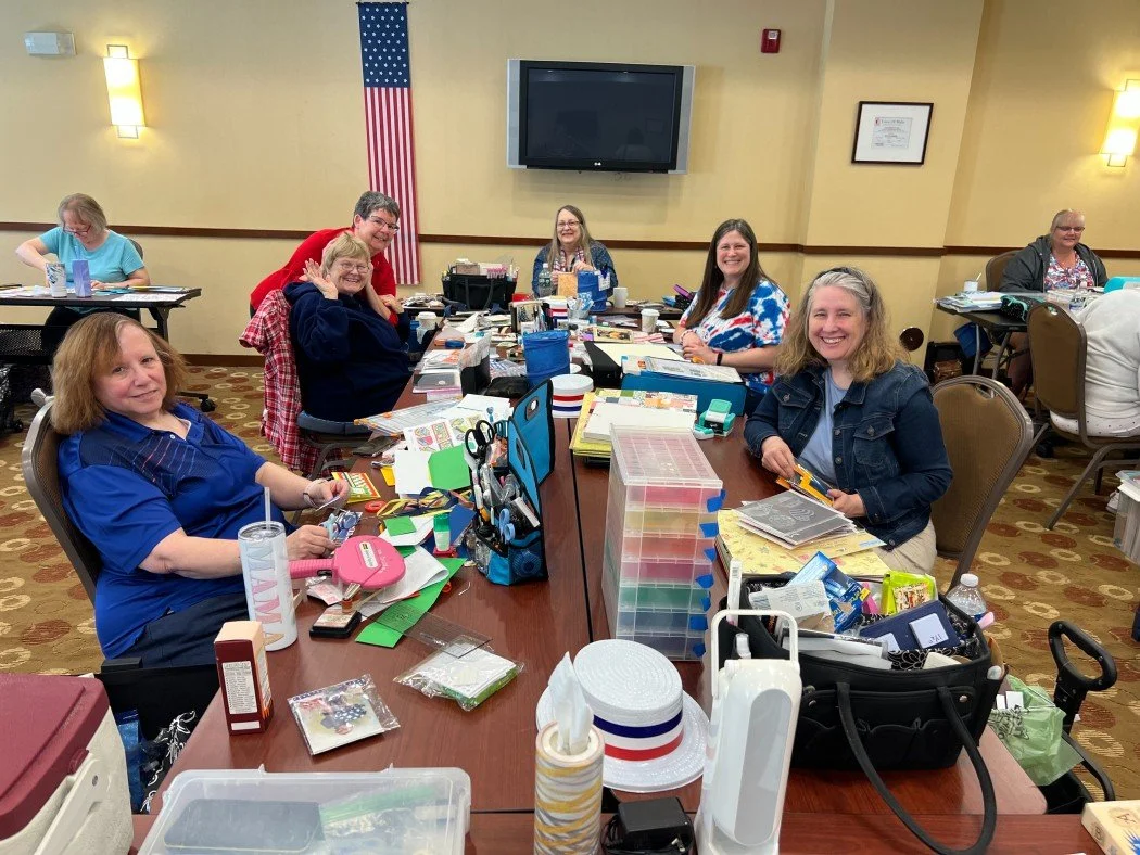 Group of women sitting around a table with crafting supplies, smiling, in a room with yellow walls, American flag, and television.