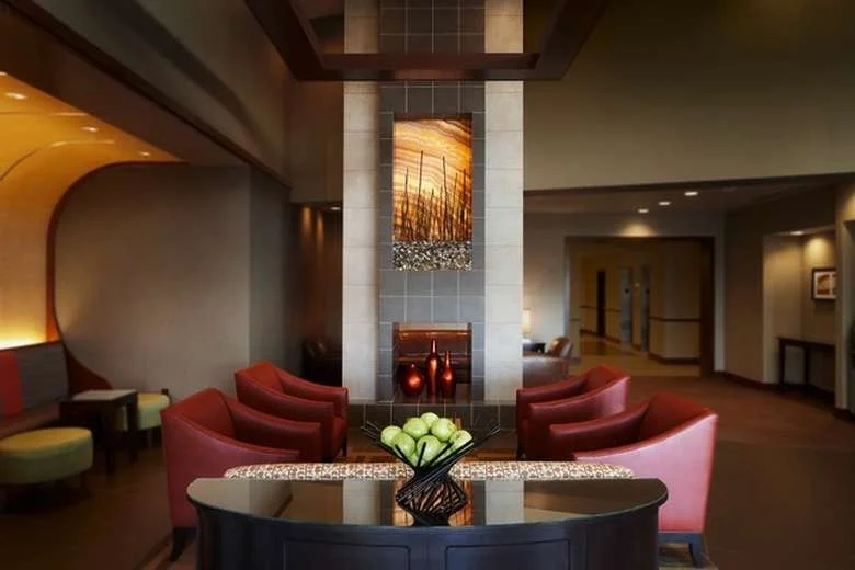 Hotel lobby with a central fireplace, surrounded by red armchairs, a dark coffee table with a bowl of green apples, and artwork above the fireplace, with warm lighting and modern decor.