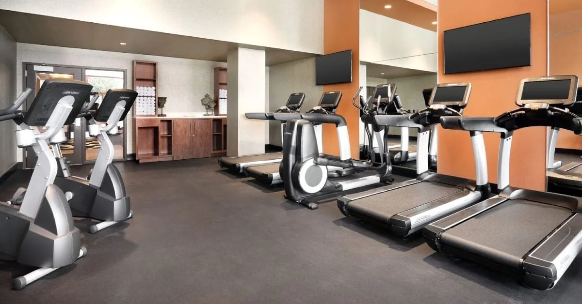 Gym with cardio equipment including treadmills, ellipticals, and stationary bikes, with wall-mounted screens and a mirror on one wall.