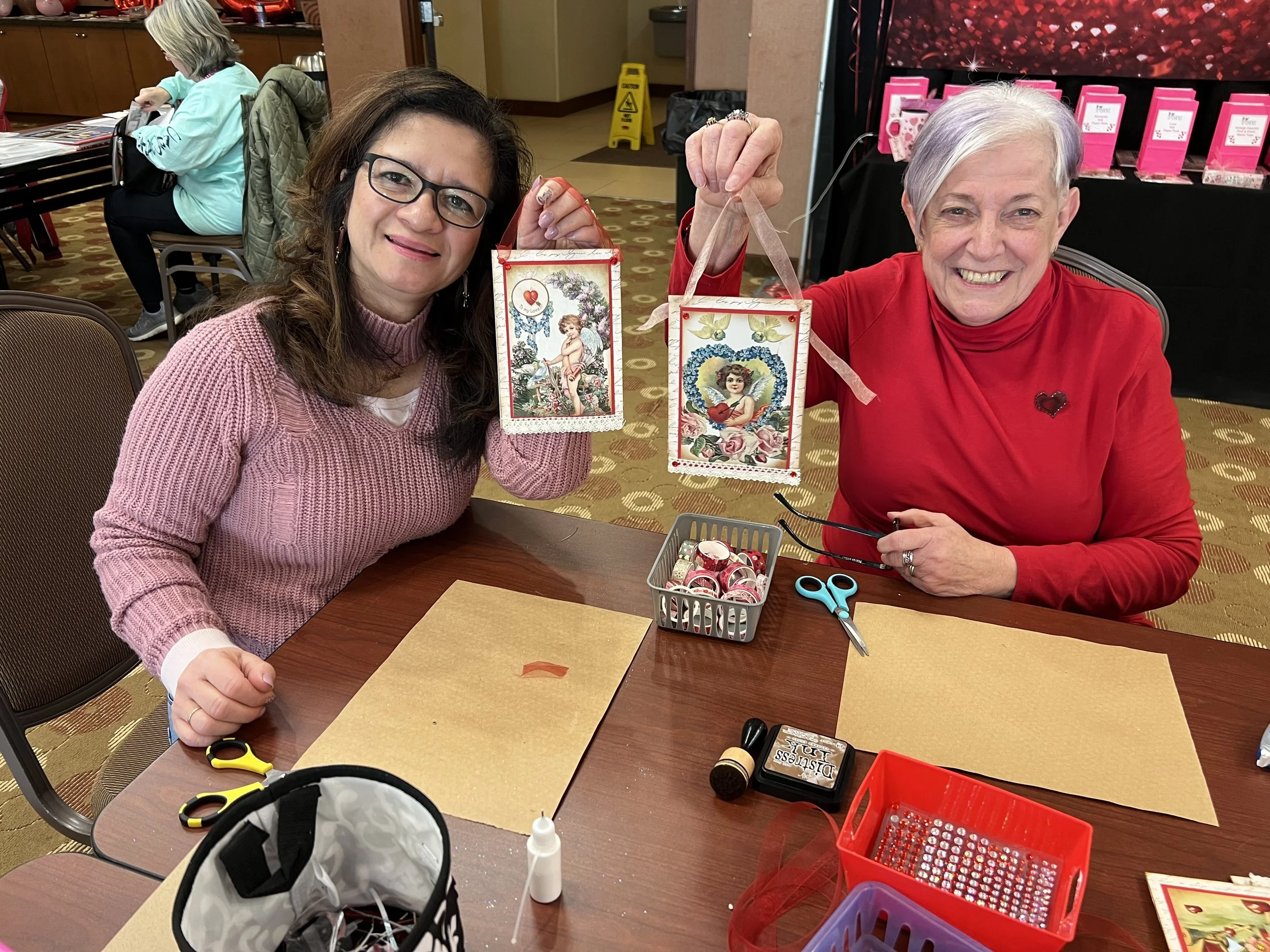 Two women sitting at a table, smiling, holding decorative Valentine's Day boxes with angel and girl illustrations. One woman is wearing a pink sweater and glasses, the other in a red top with a heart pin. The table has craft supplies and a pair of sc