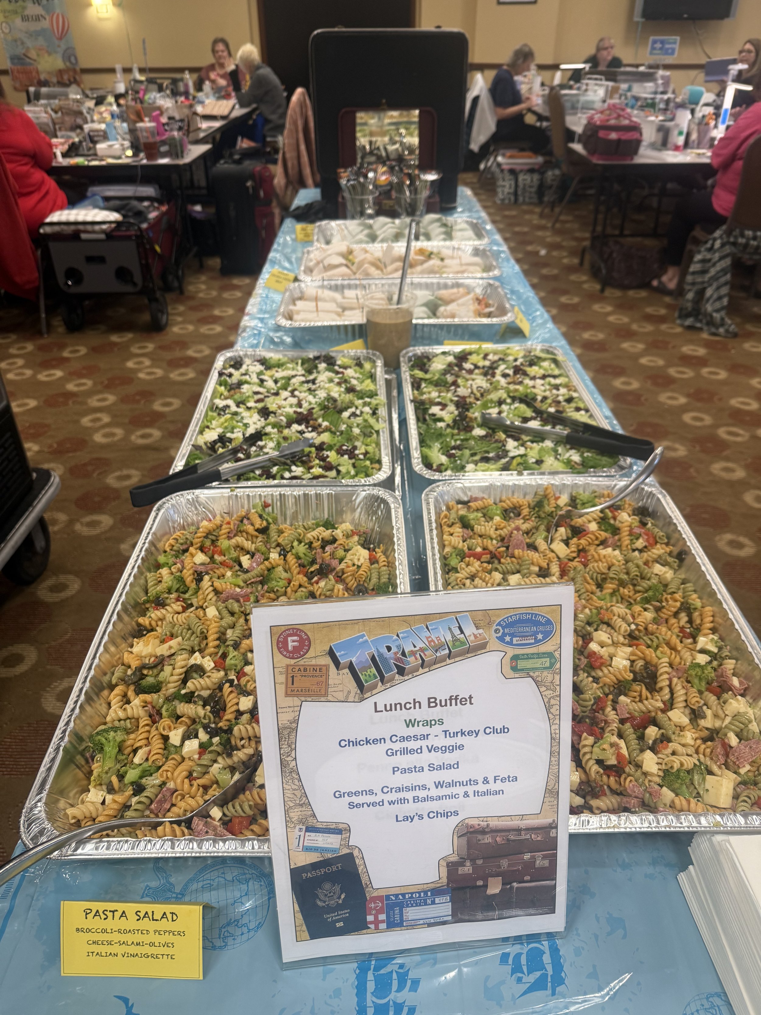 A lunch buffet table with various pasta salads, greens, and wraps at a conference room. In the background, people are seated at tables, some eating and talking.