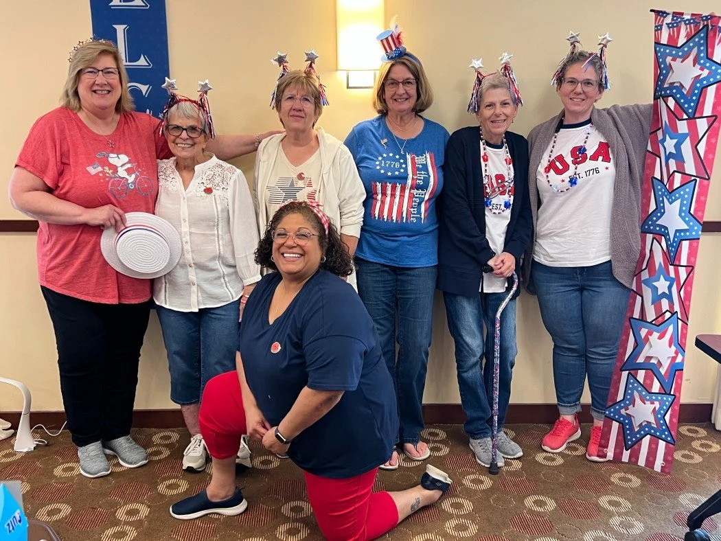 A group of seven women celebrating Independence Day, wearing patriotic accessories like star headbands and shirts with USA motifs. They are smiling and posing together in a decorated room.